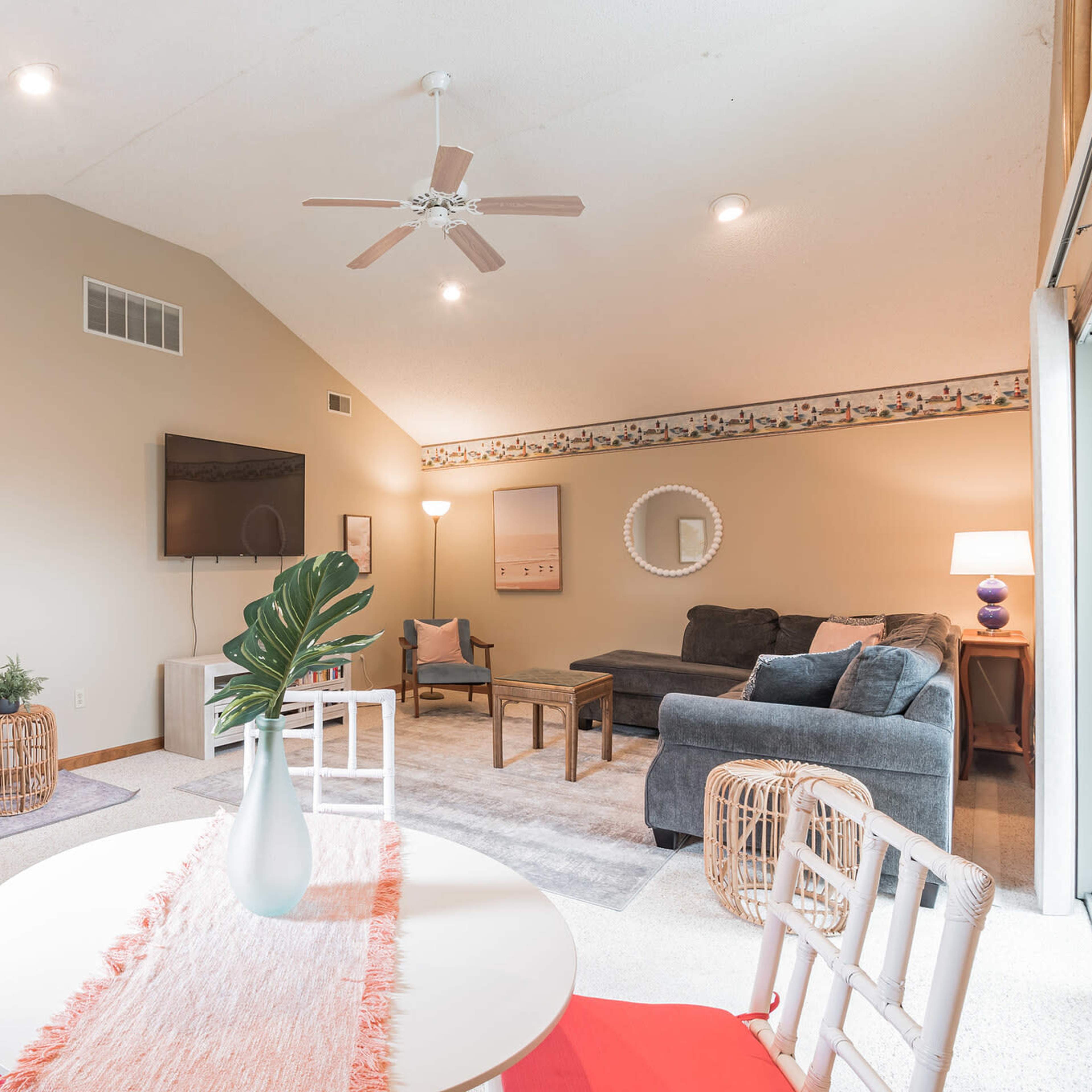A bright living area with a vaulted ceiling, featuring a dining table with red chairs, a plush sofa, a TV mounted on the wall, and decorative elements.