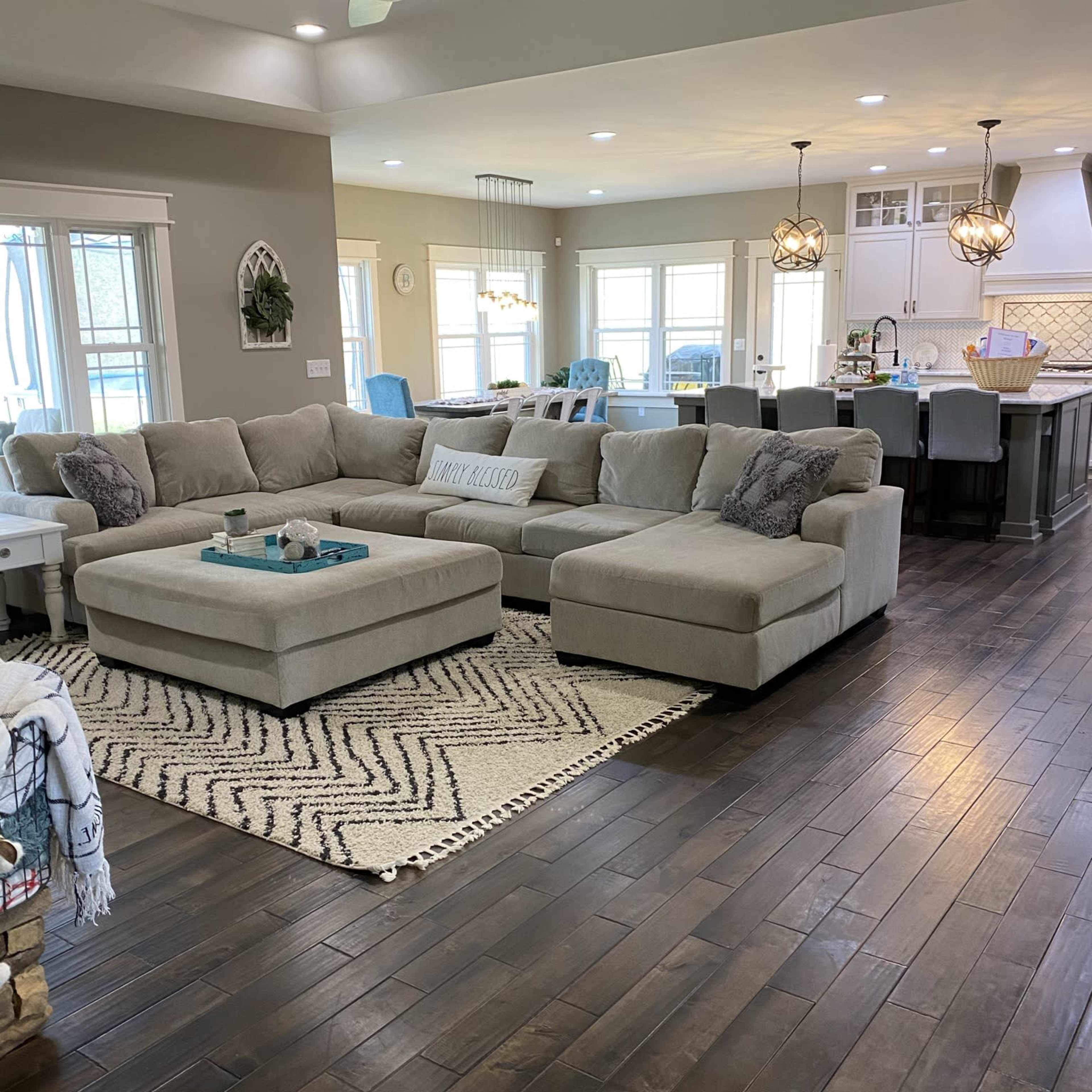 A spacious living room with a large beige sectional sofa and a patterned area rug, leading to an open kitchen area with modern fixtures and large windows.