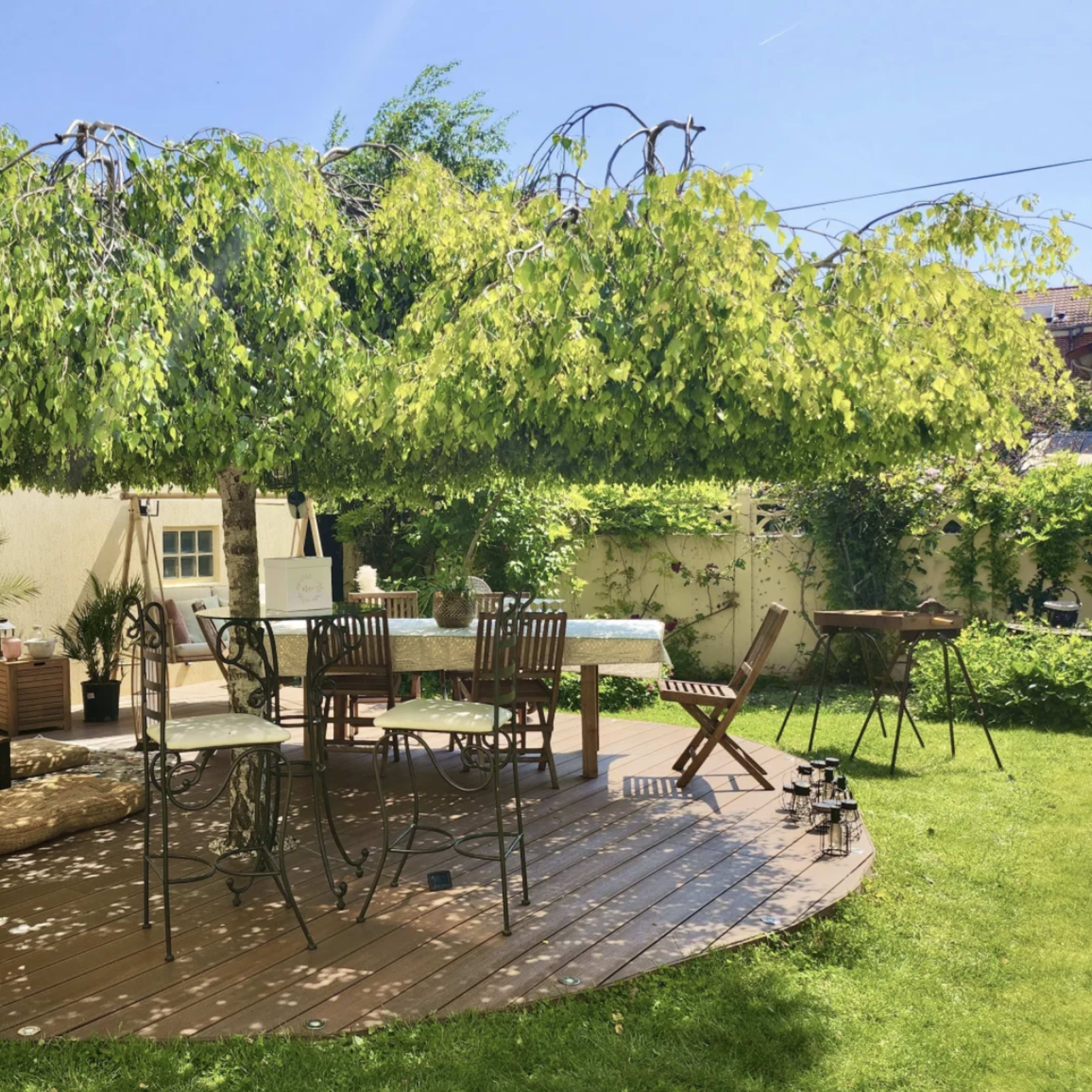 A shaded outdoor seating area features a wooden deck surrounded by greenery, with a table and chairs set for dining.