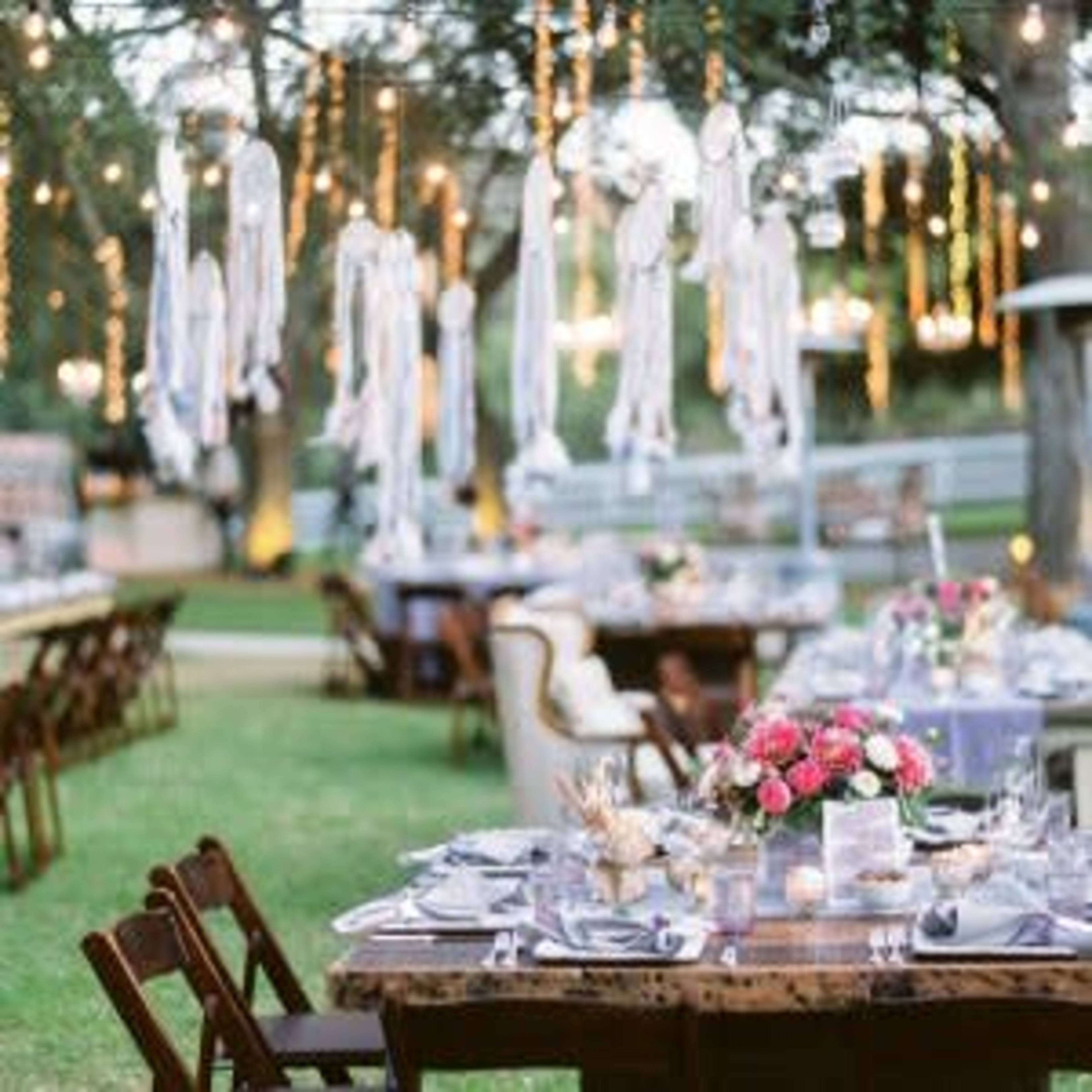 An outdoor dining setup is arranged with tables adorned with floral centerpieces and hanging lights, creating an elegant atmosphere.