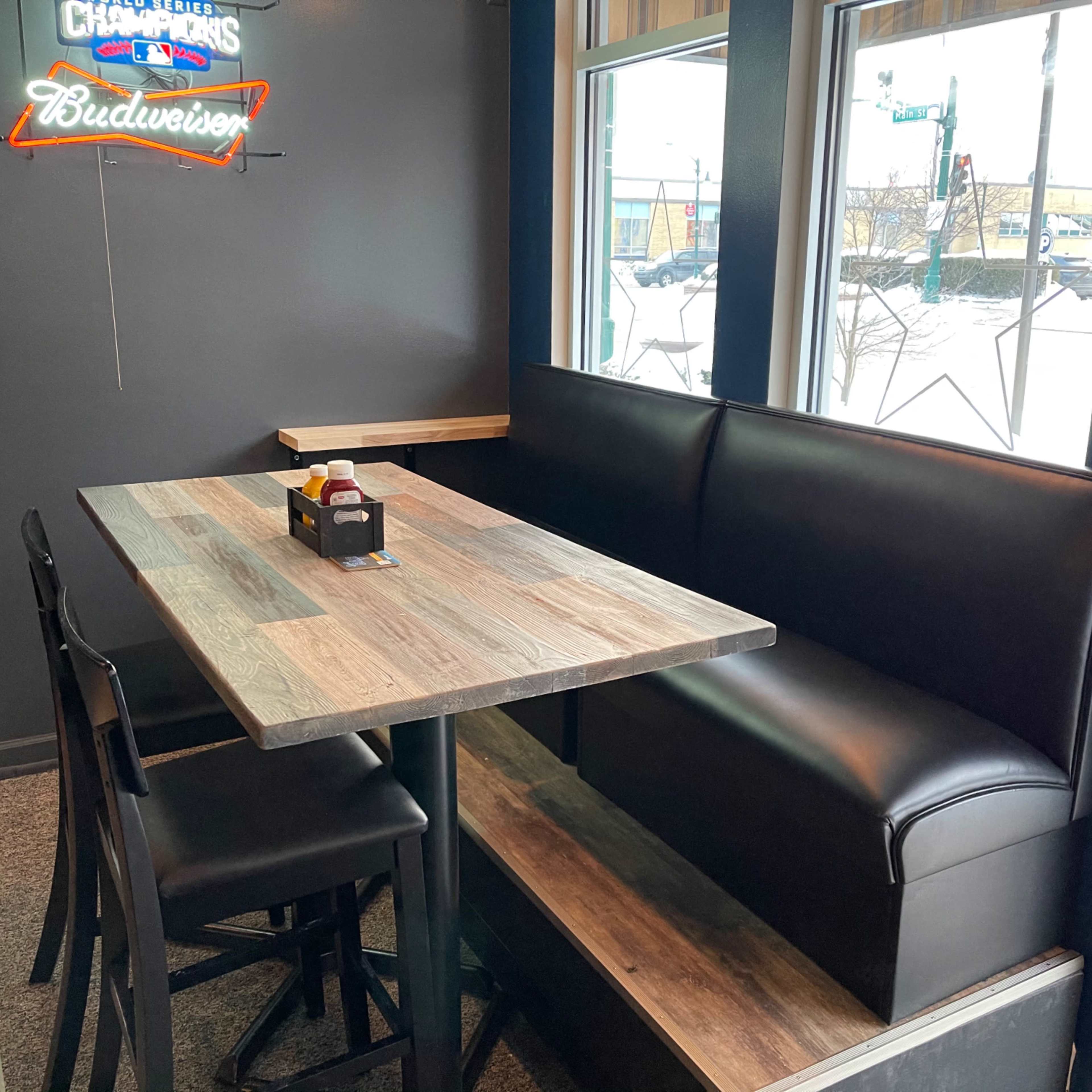 A wooden table with black cushioned seating is positioned next to large windows in a dining area.