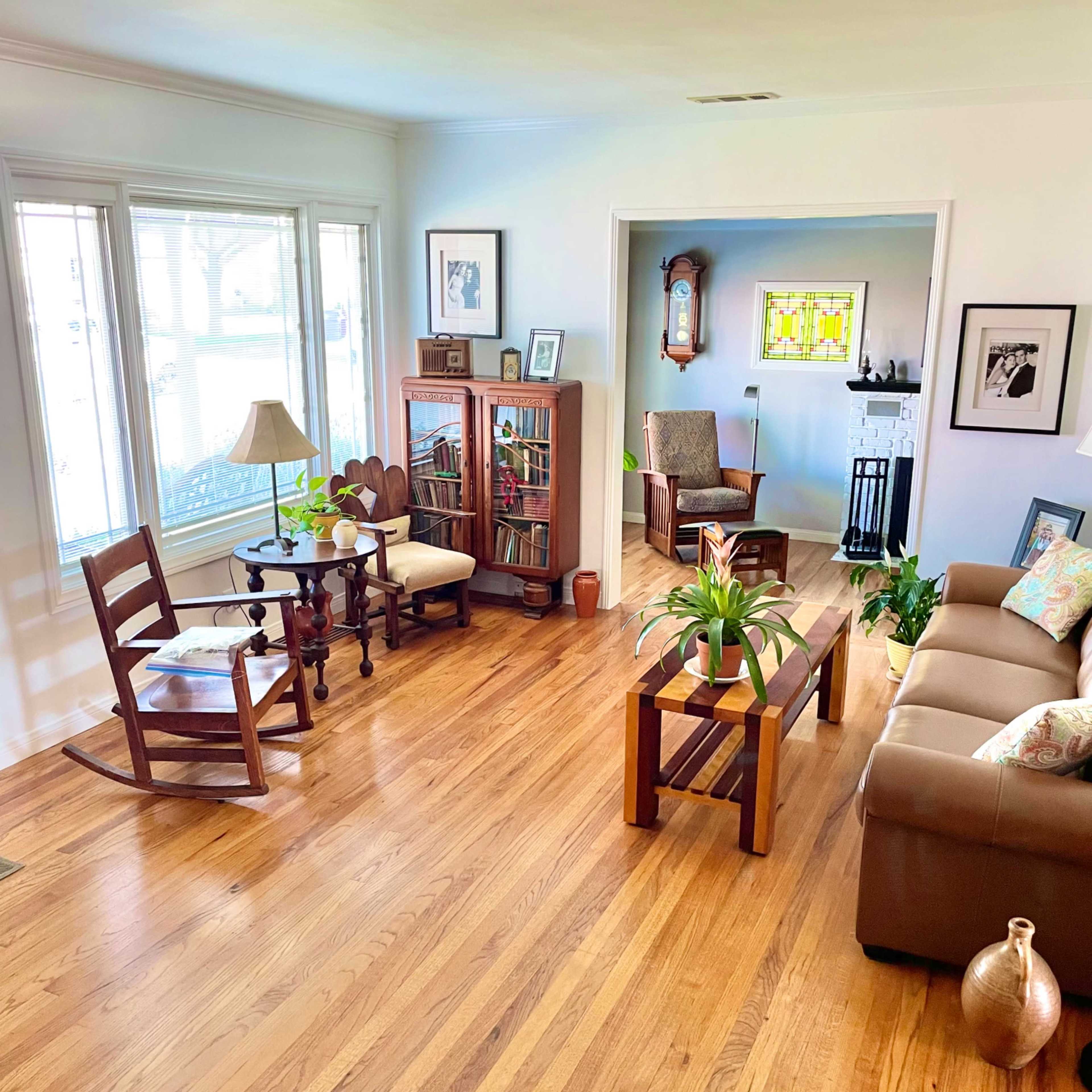 A brightly lit living room with wooden floors, a seating area, a rocking chair, a bookshelf, and plants.
