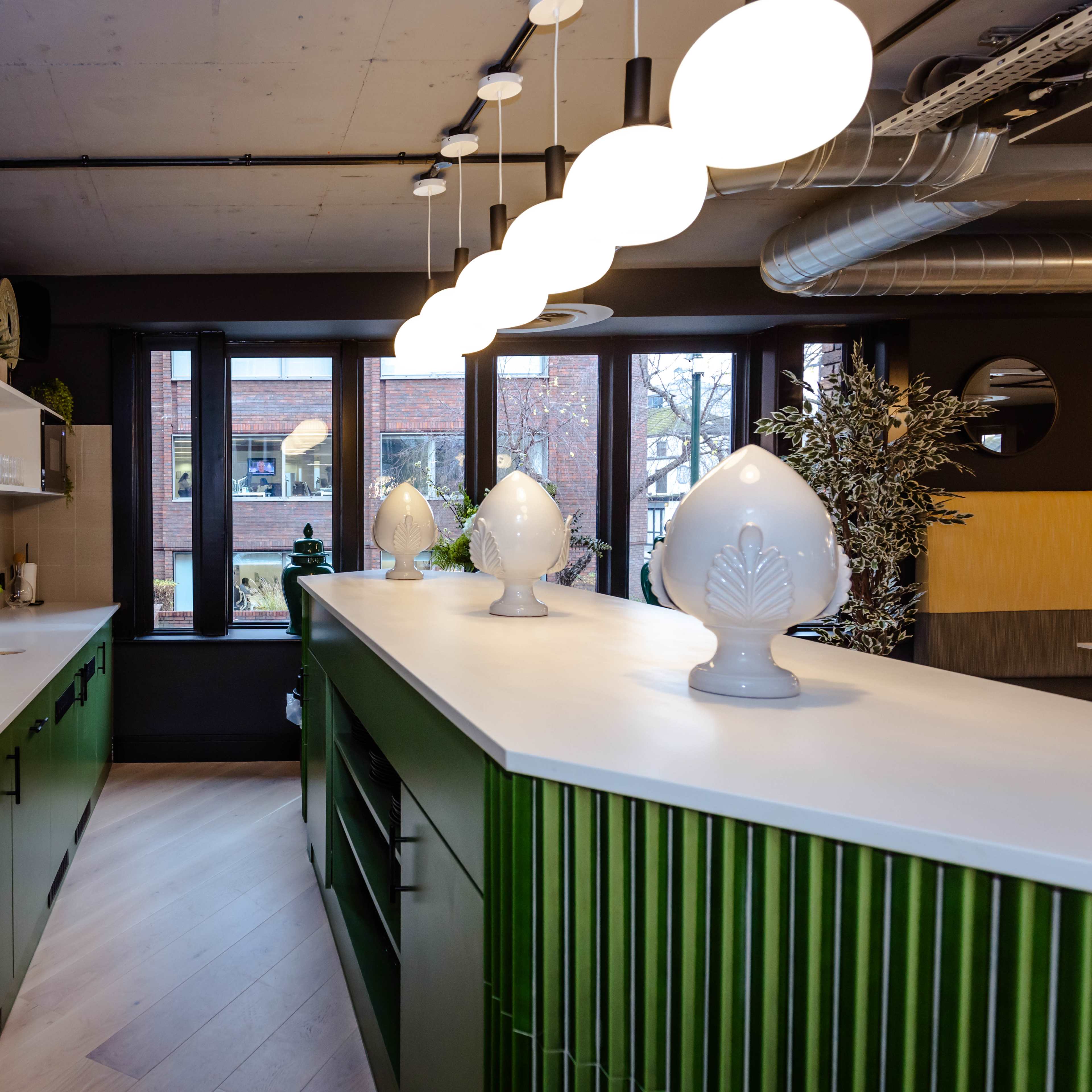 A modern kitchen with green cabinetry, a white countertop, and decorative white vases, illuminated by circular pendant lights.