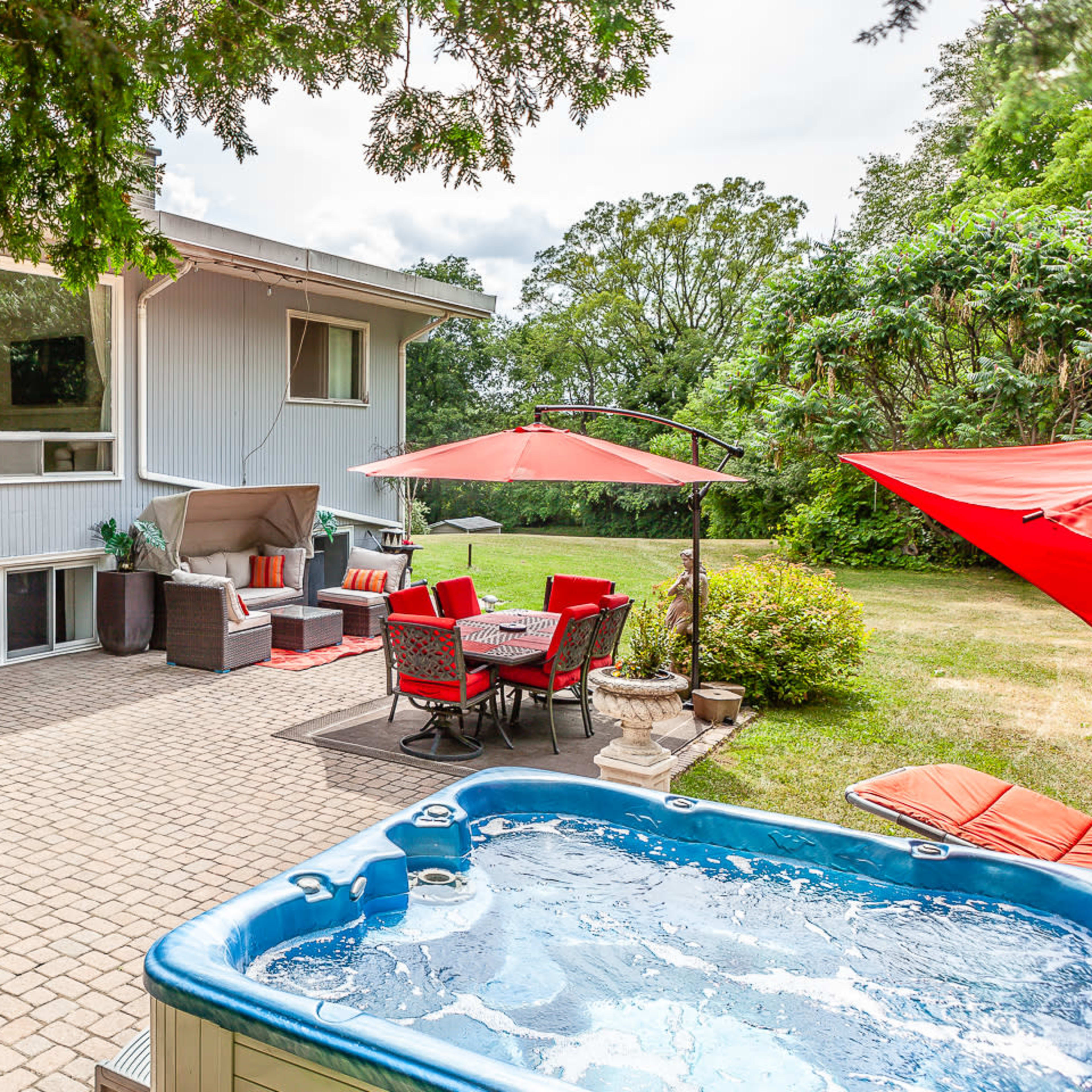 A backyard patio with a hot tub, seating area, and a dining set under red umbrellas, surrounded by greenery.