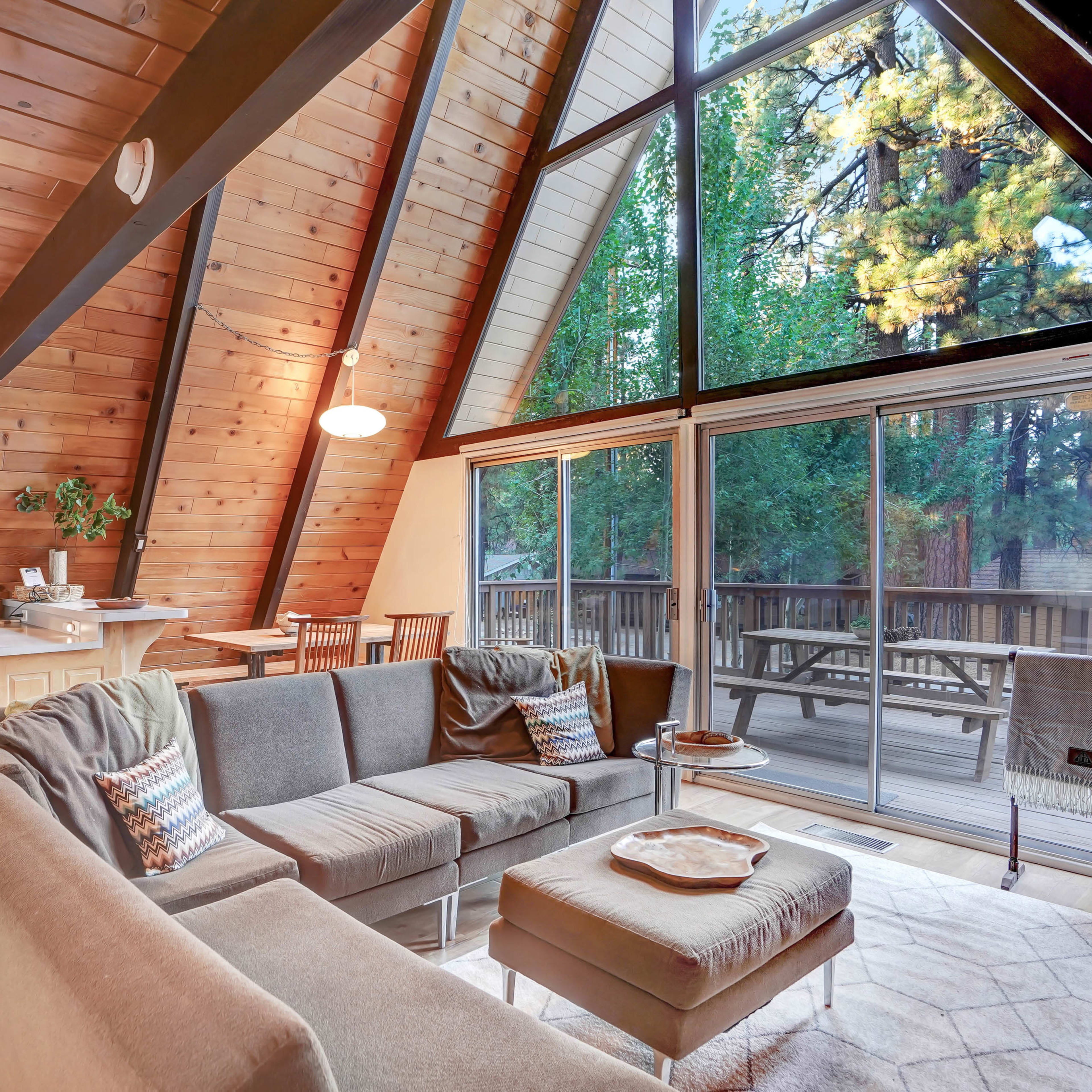 A modern A-frame cabin interior with a wooden kitchen and a spacious living area featuring a sectional sofa and large windows overlooking a deck and trees.