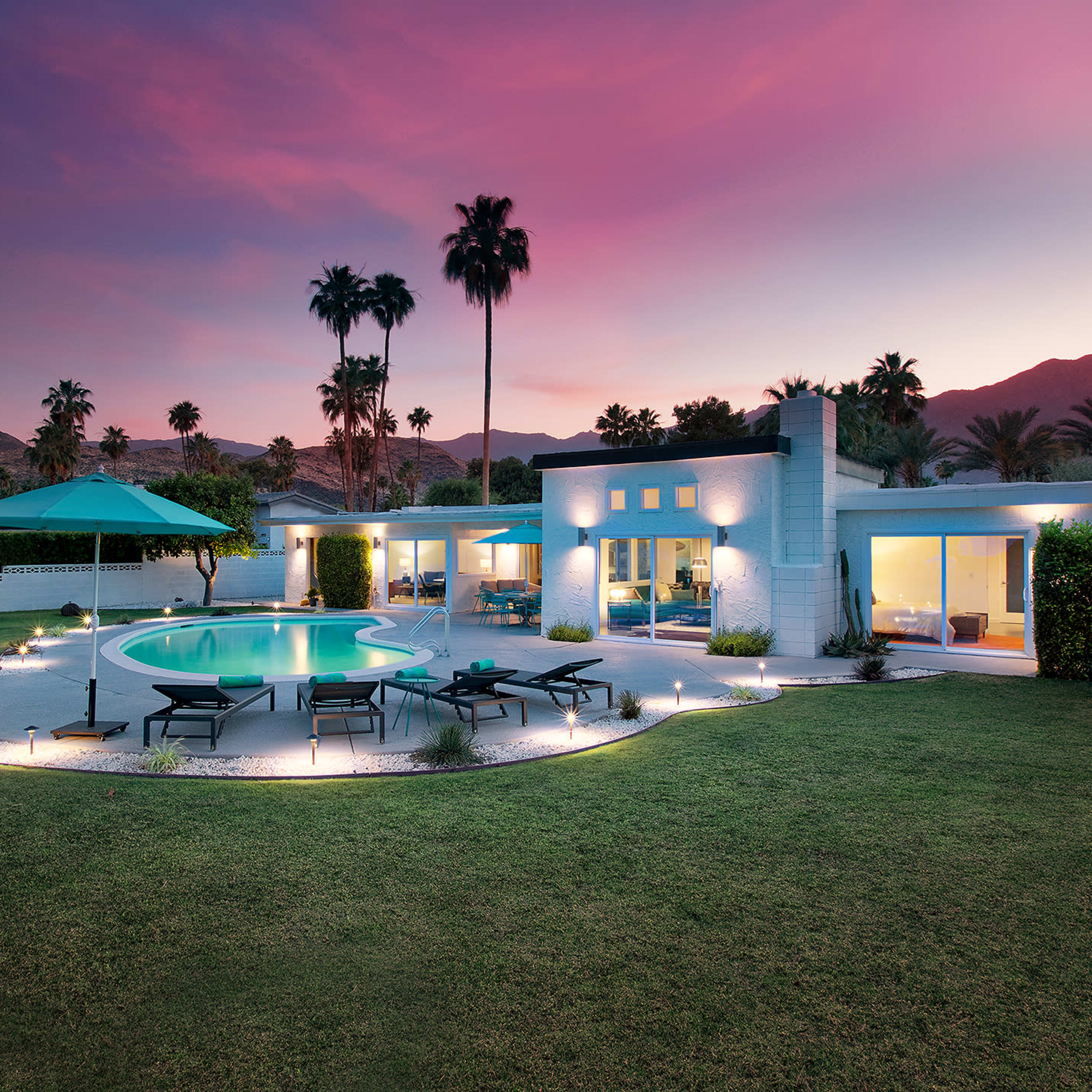 A modern house with a swimming pool and palm trees is set against a colorful sunset in a landscaped yard.