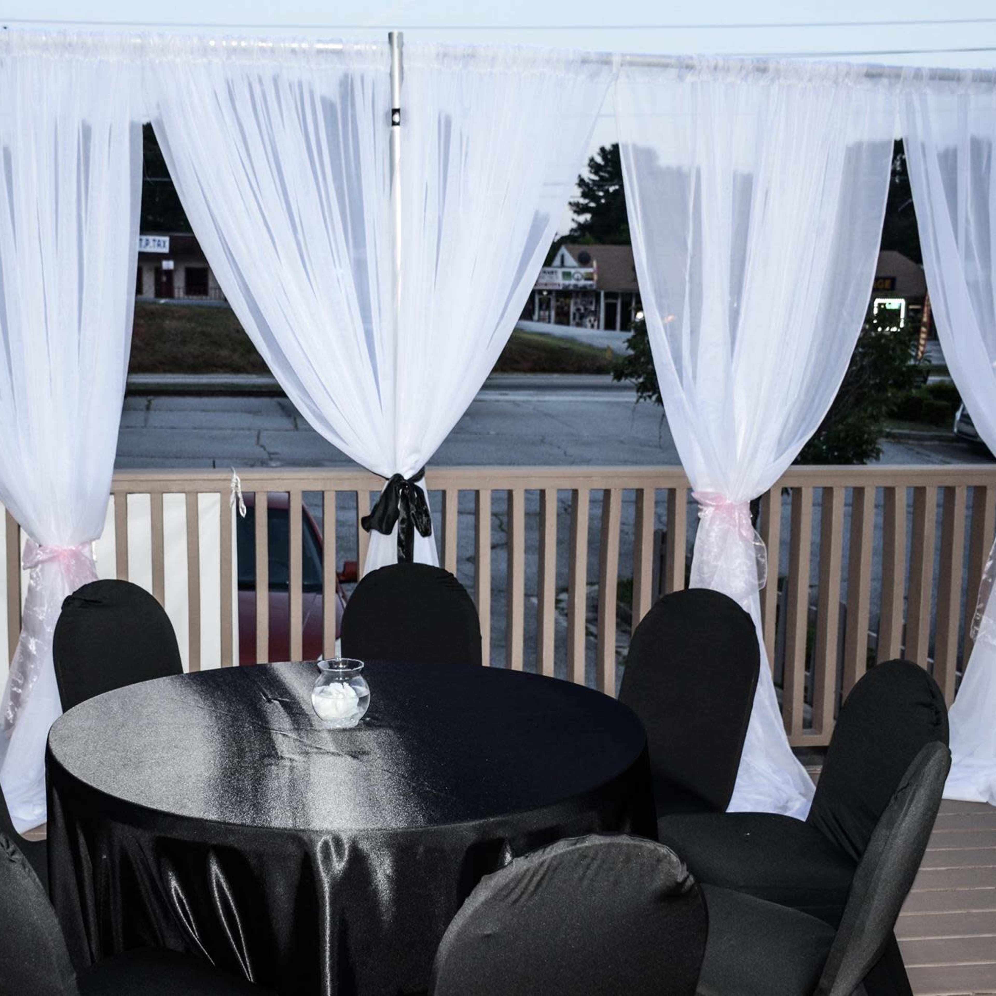 A round black table surrounded by black chairs is positioned on a deck, with sheer white curtains draping from a railing.