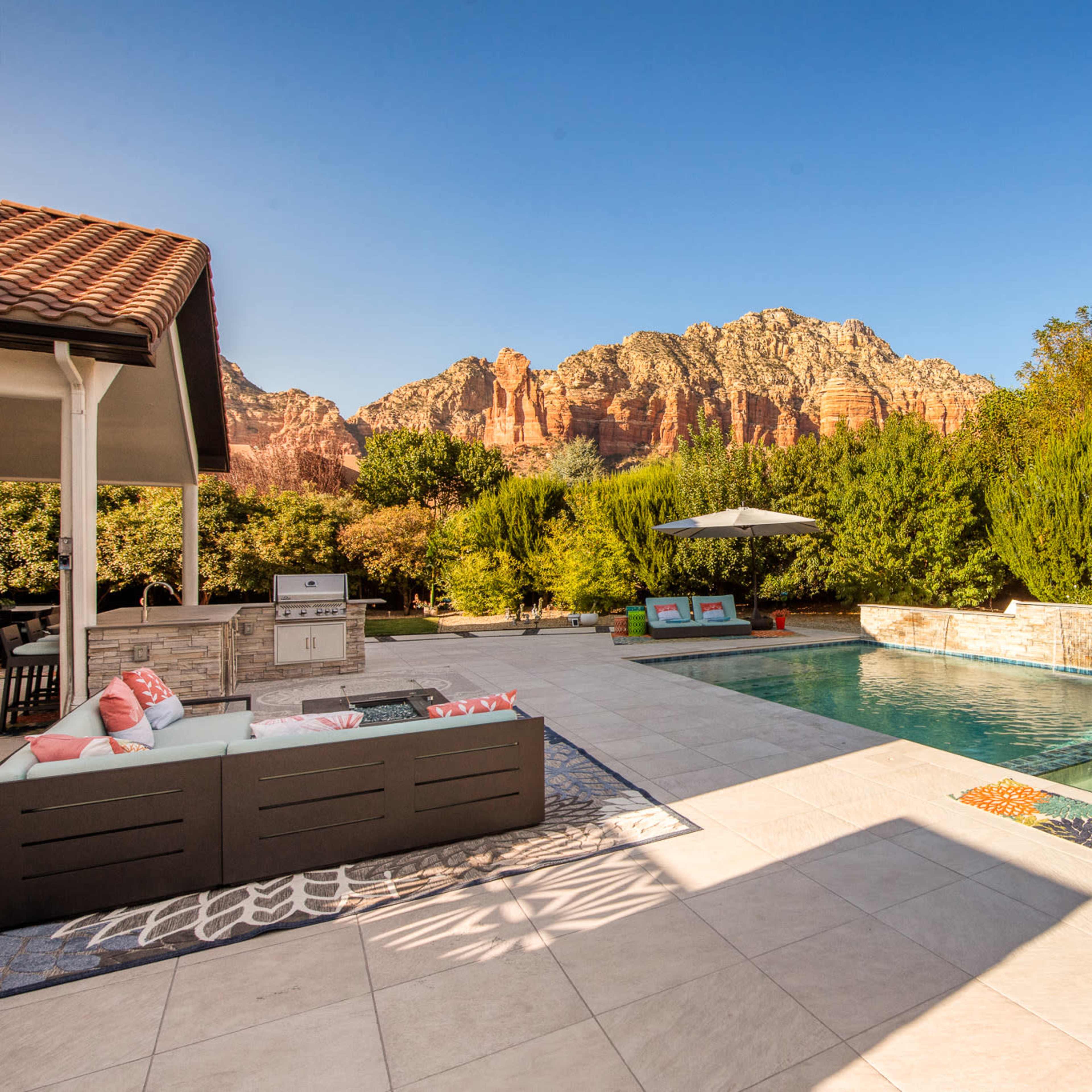 A backyard features a swimming pool and spa surrounded by stone landscaping and seating areas, with mountains in the background.