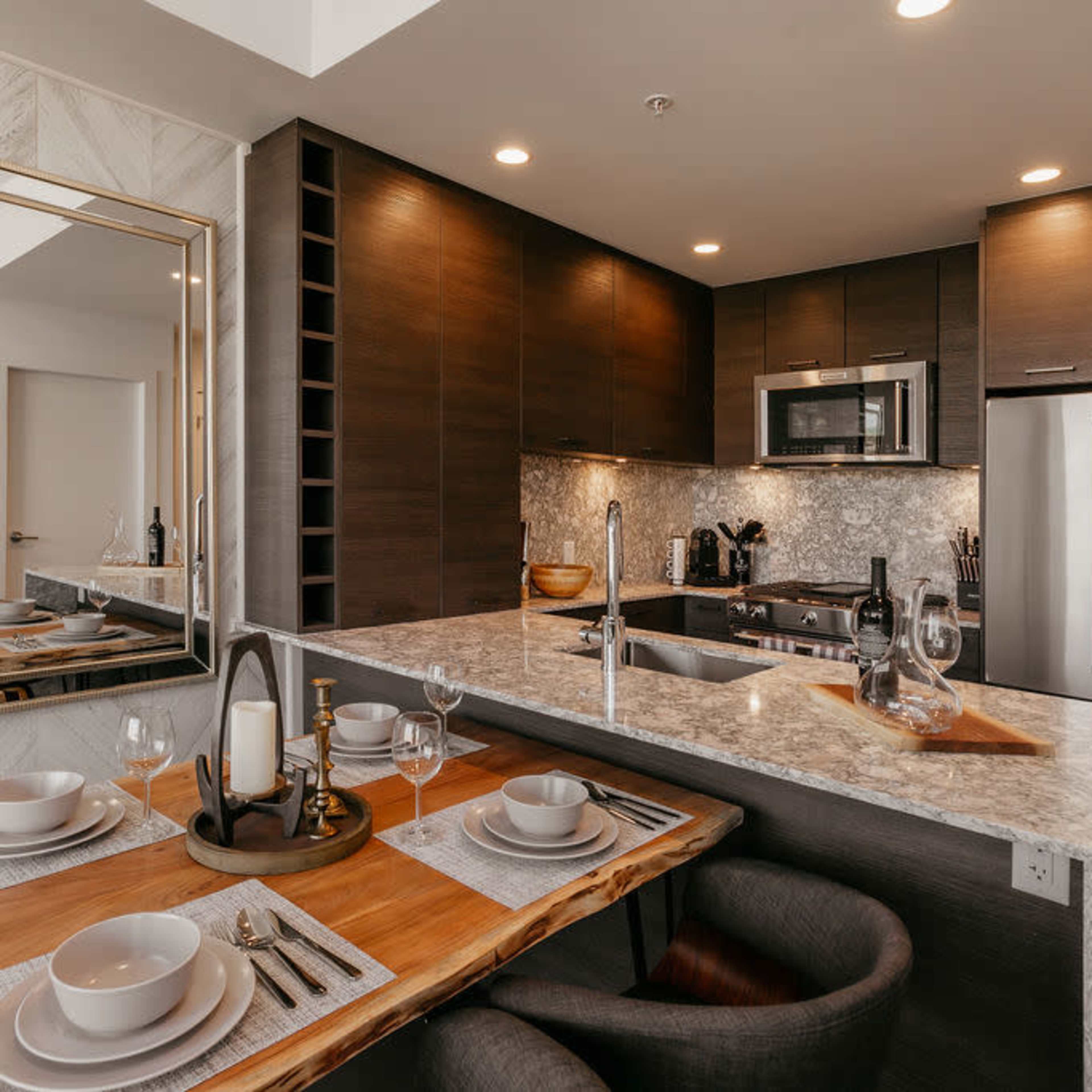 A modern kitchen and dining area featuring a marble countertop, dark cabinetry, and a large mirror reflecting the dining table set with plates and glasses.
