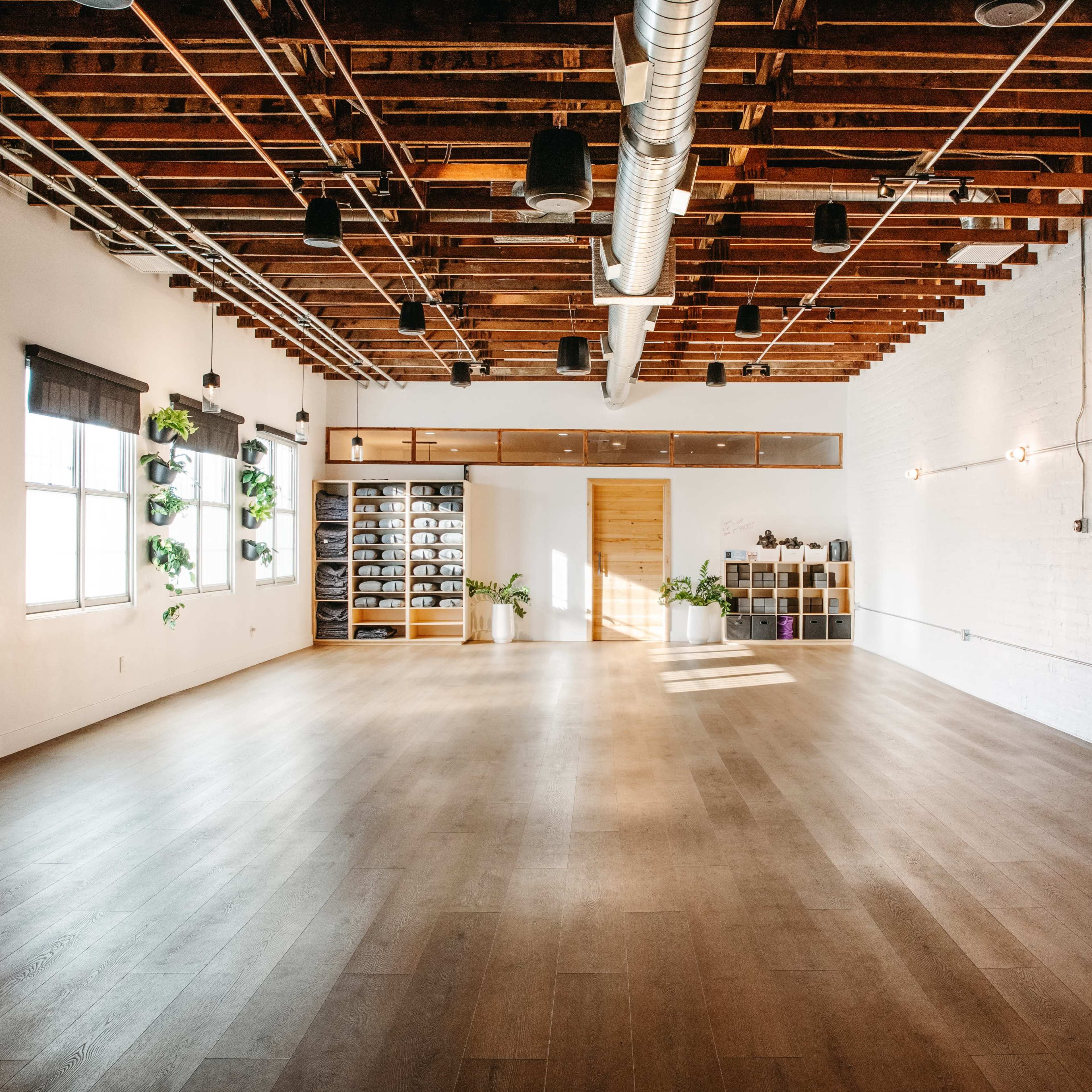 A spacious, well-lit studio with wooden flooring, exposed wooden beams, and minimalistic decor, featuring plants and shelving against the walls.