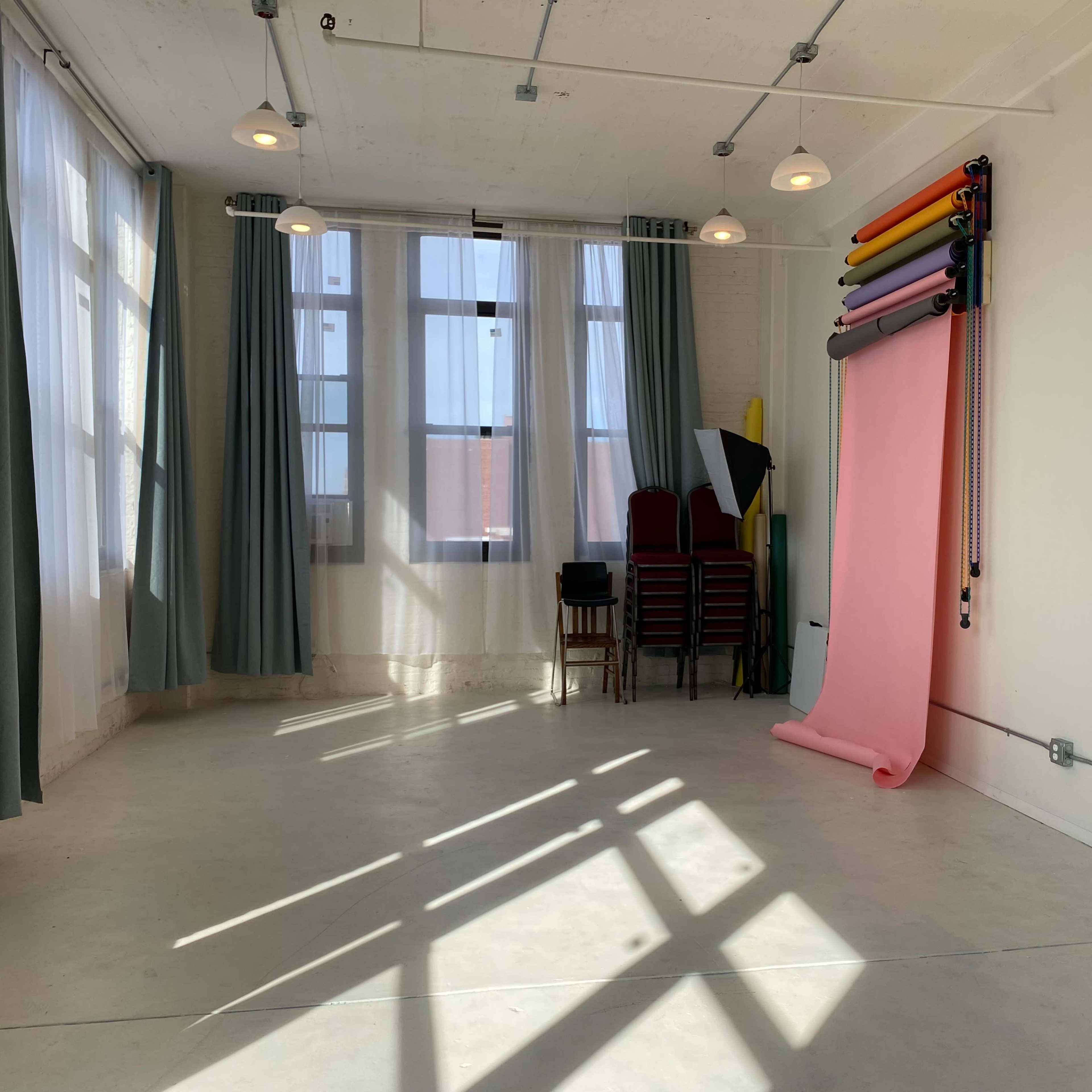 A bright room with large windows, green curtains, a stack of chairs, and a roll of photography backdrop paper in various colors.