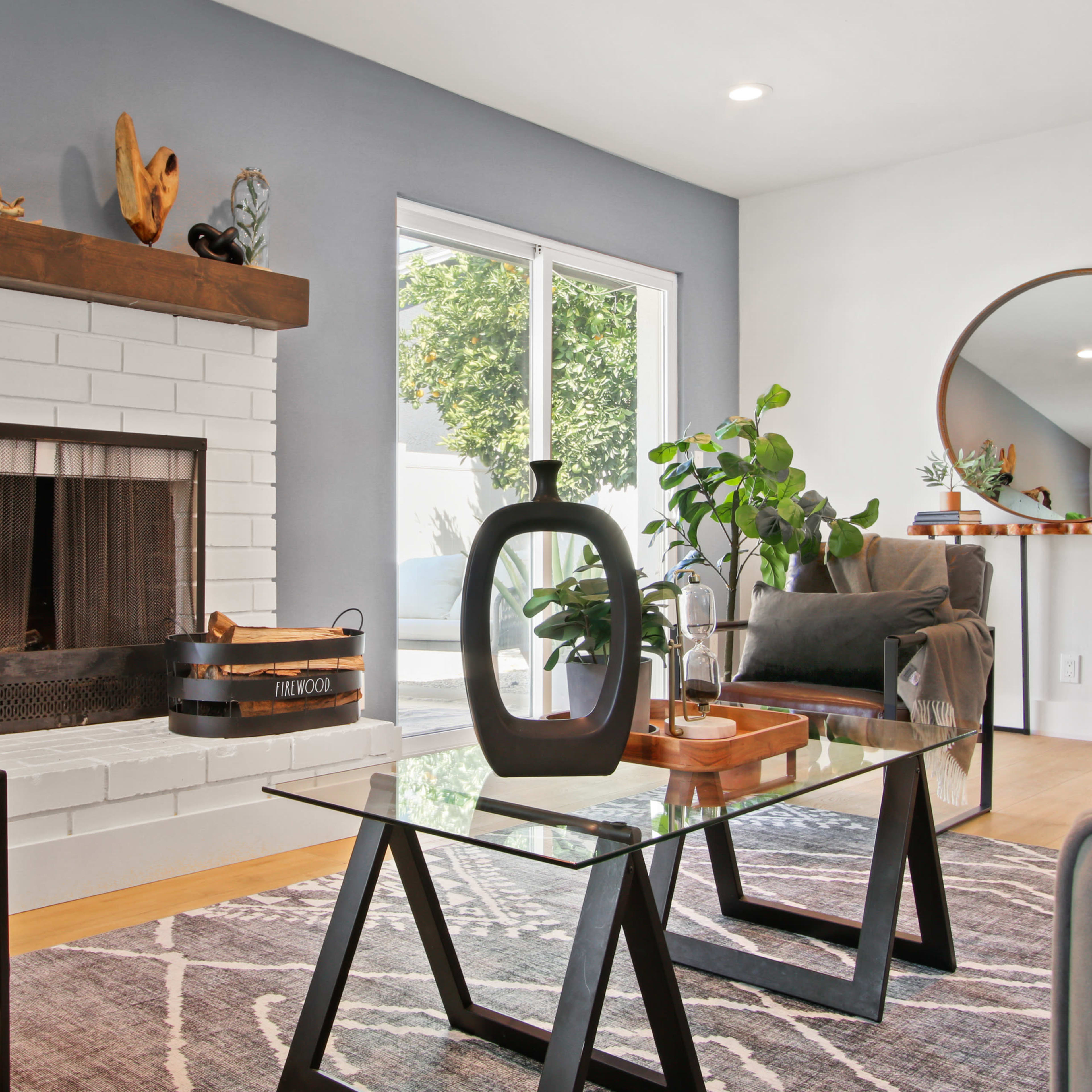 A modern living room with a white brick fireplace, a glass coffee table, and a seating area featuring two armchairs and a large window bringing in natural light.