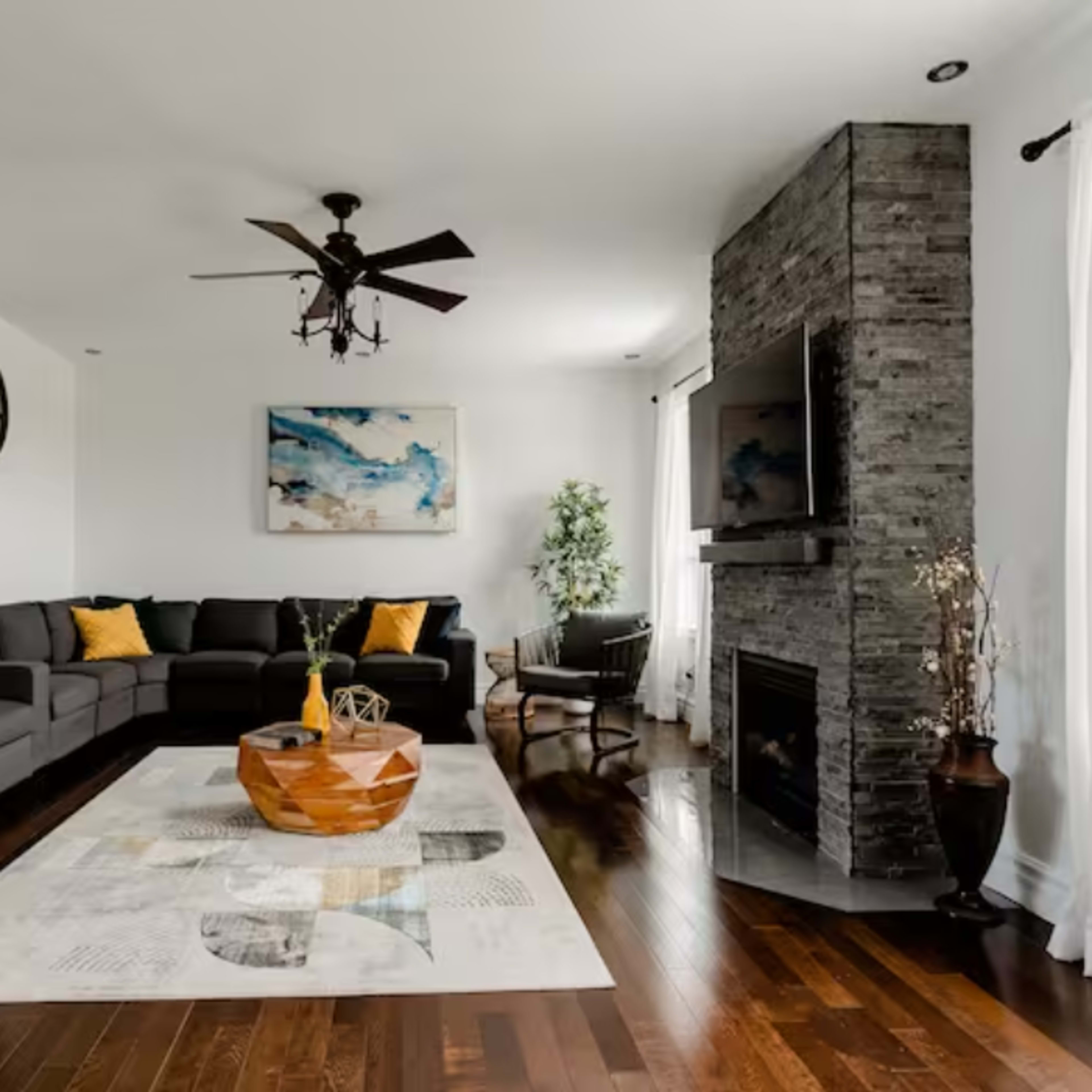 A modern living room featuring a sectional sofa, a stone fireplace, a large area rug, and a ceiling fan, with decorative plants and artwork on the walls.