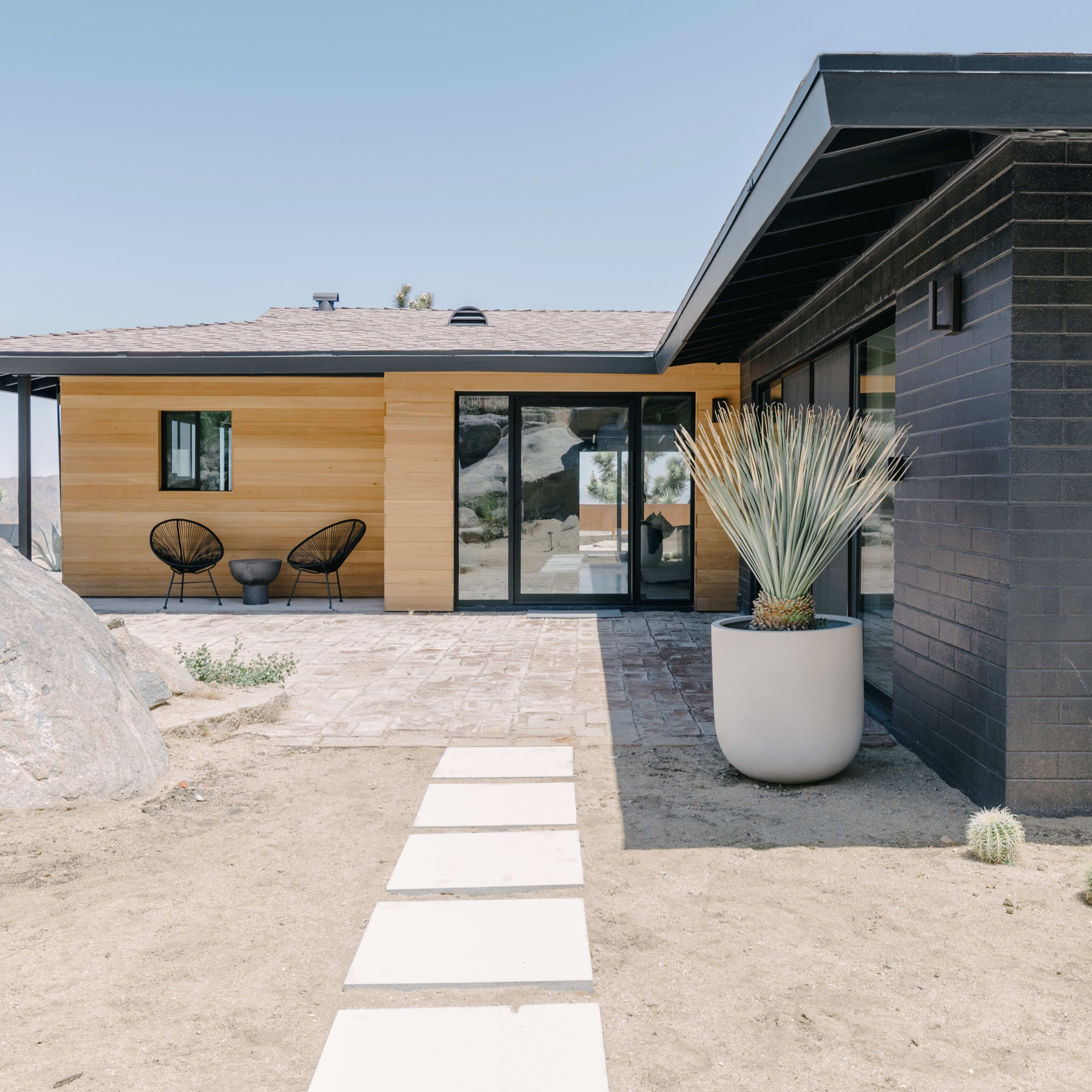 A modern house with a combination of wood and black brick exteriors, featuring a landscaped yard with a stone pathway and a large potted plant.