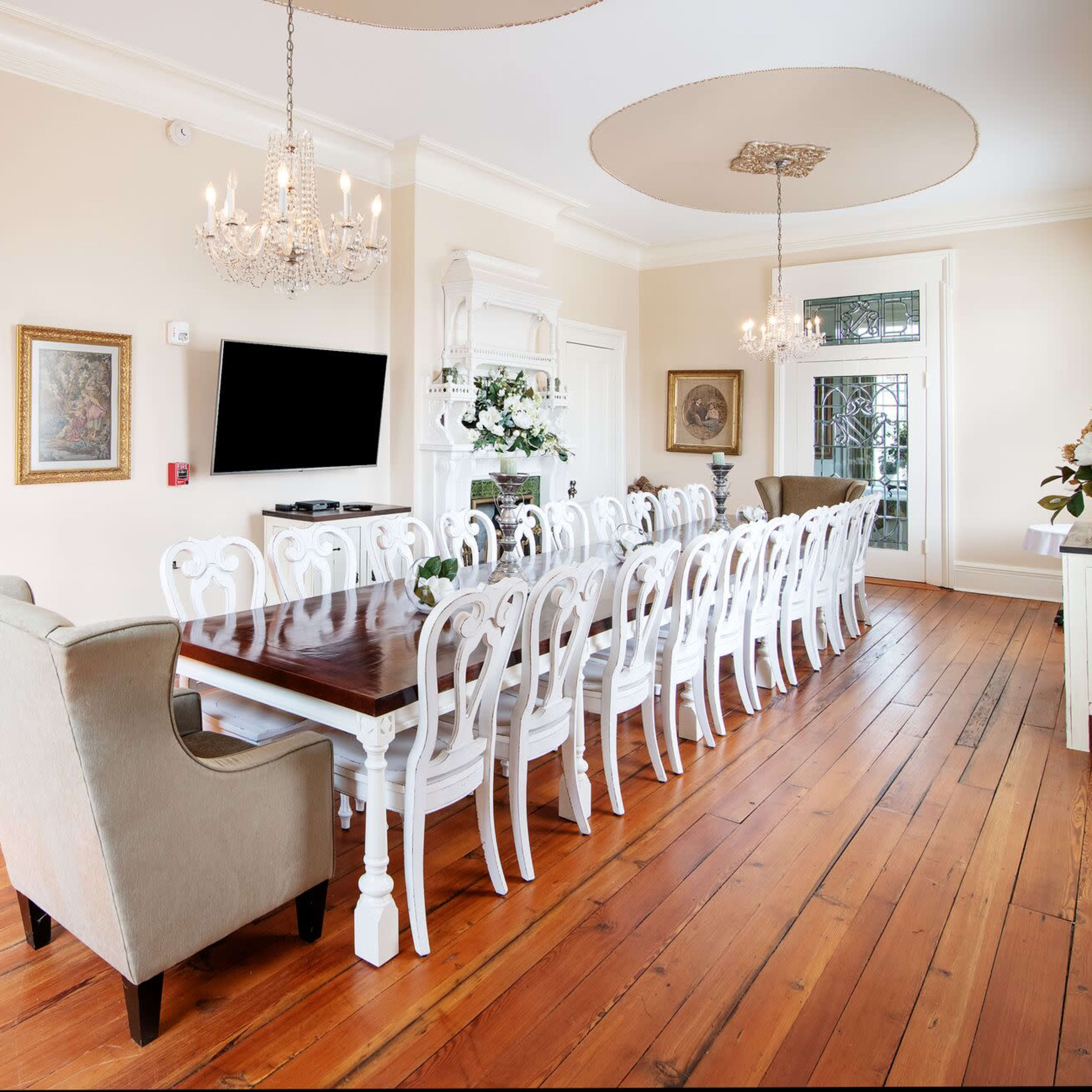 A spacious dining room features a long wooden table surrounded by white chairs, with a chandelier overhead and an exit sign on the wall.