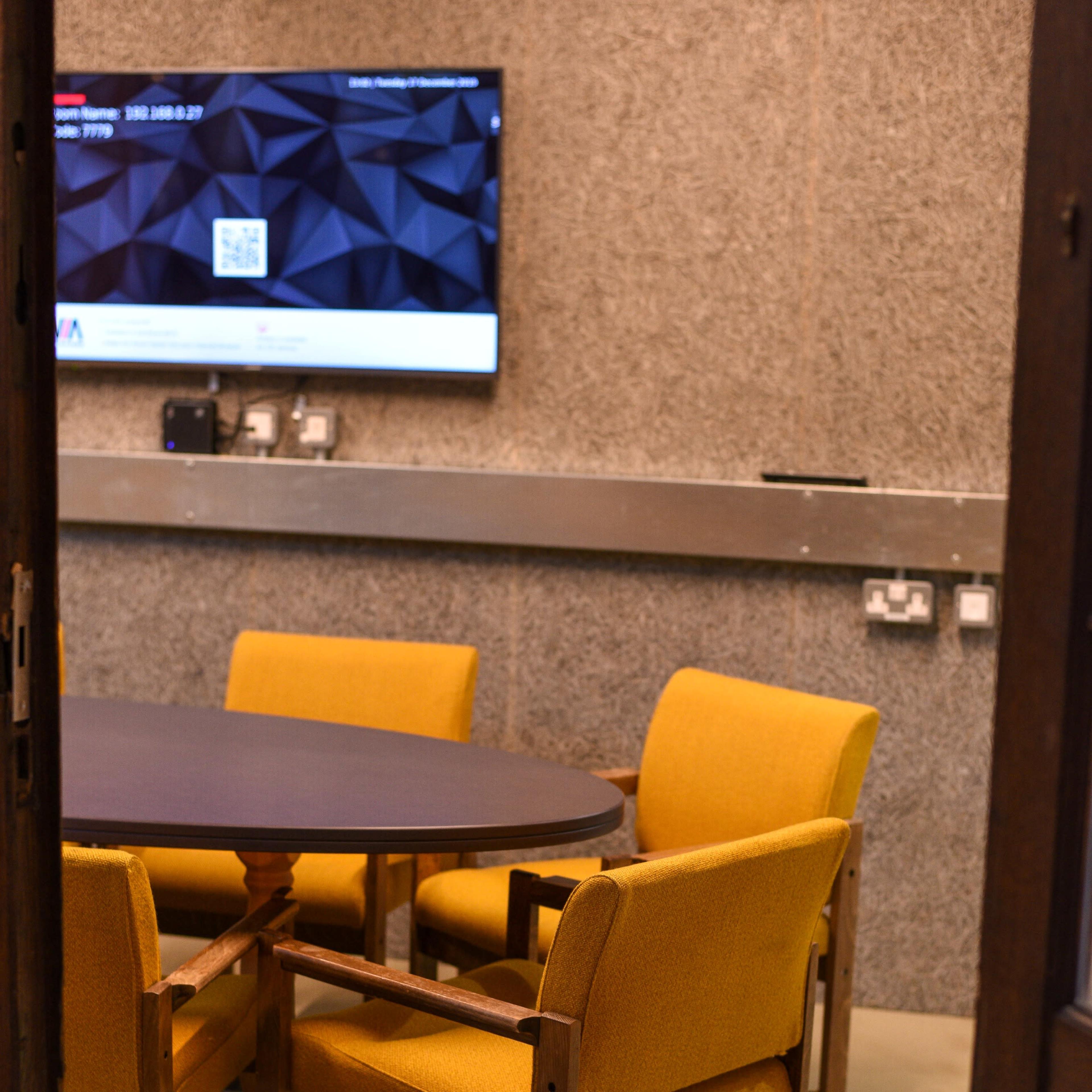 A small meeting room features a round table surrounded by four yellow armchairs, with a large screen mounted on the wall.