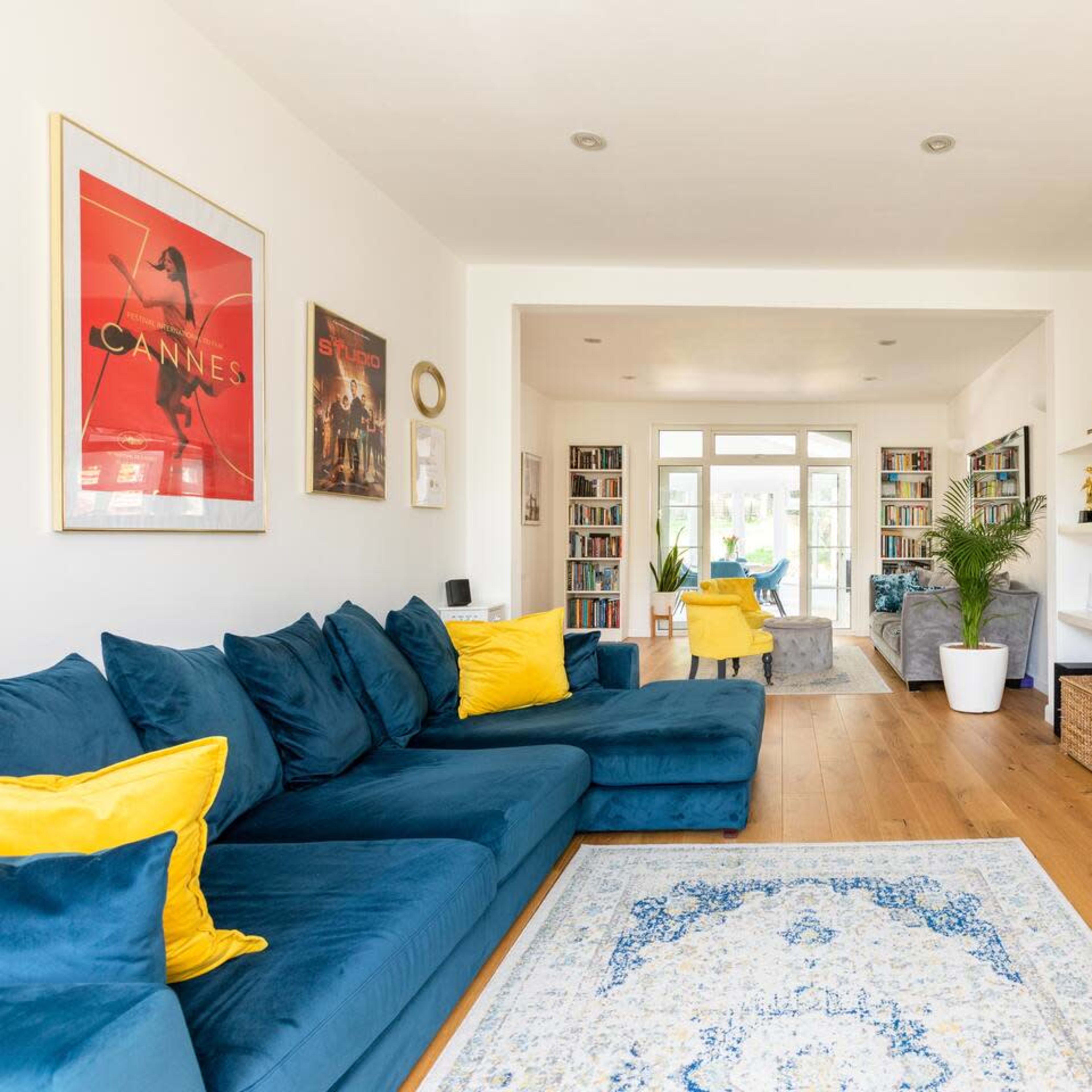 The living room features a blue sectional sofa with yellow cushions, a large television mounted on the wall, and shelves filled with books.