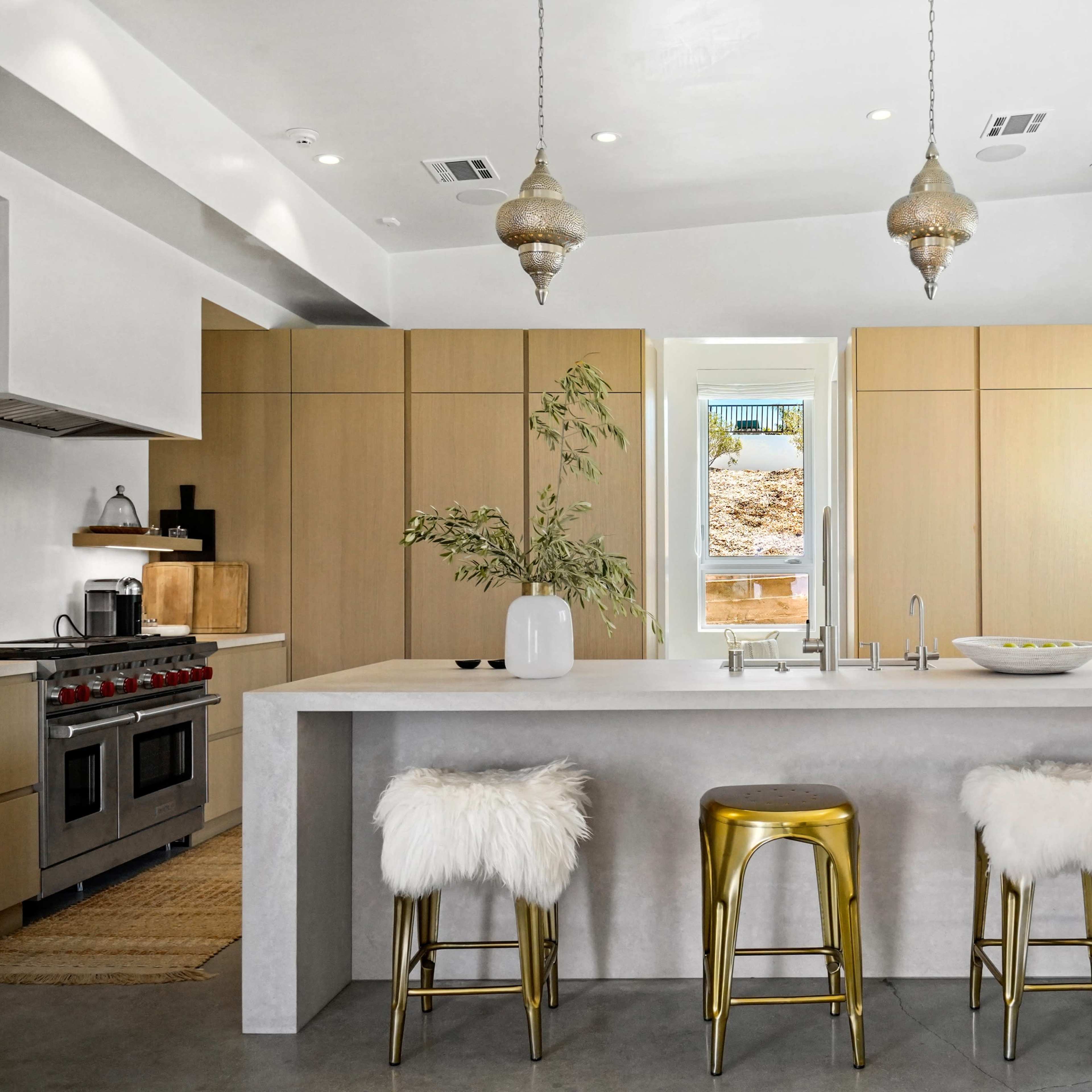 A modern kitchen features a concrete island with gold stools and wooden cabinetry, illuminated by pendant lights.