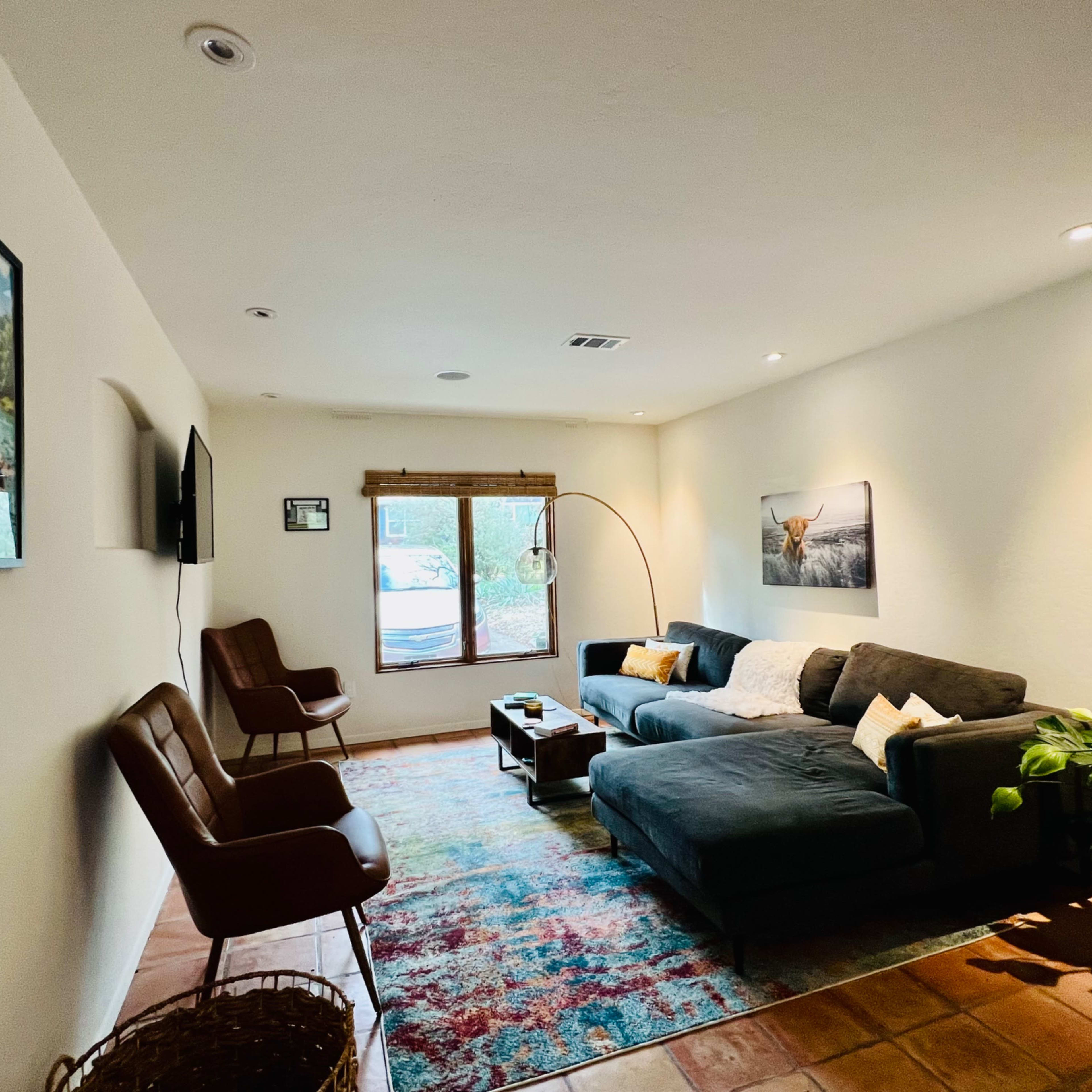 A living room features a gray sectional sofa, two brown chairs, a floor lamp, and a colorful area rug, with wall art and a window letting in natural light.