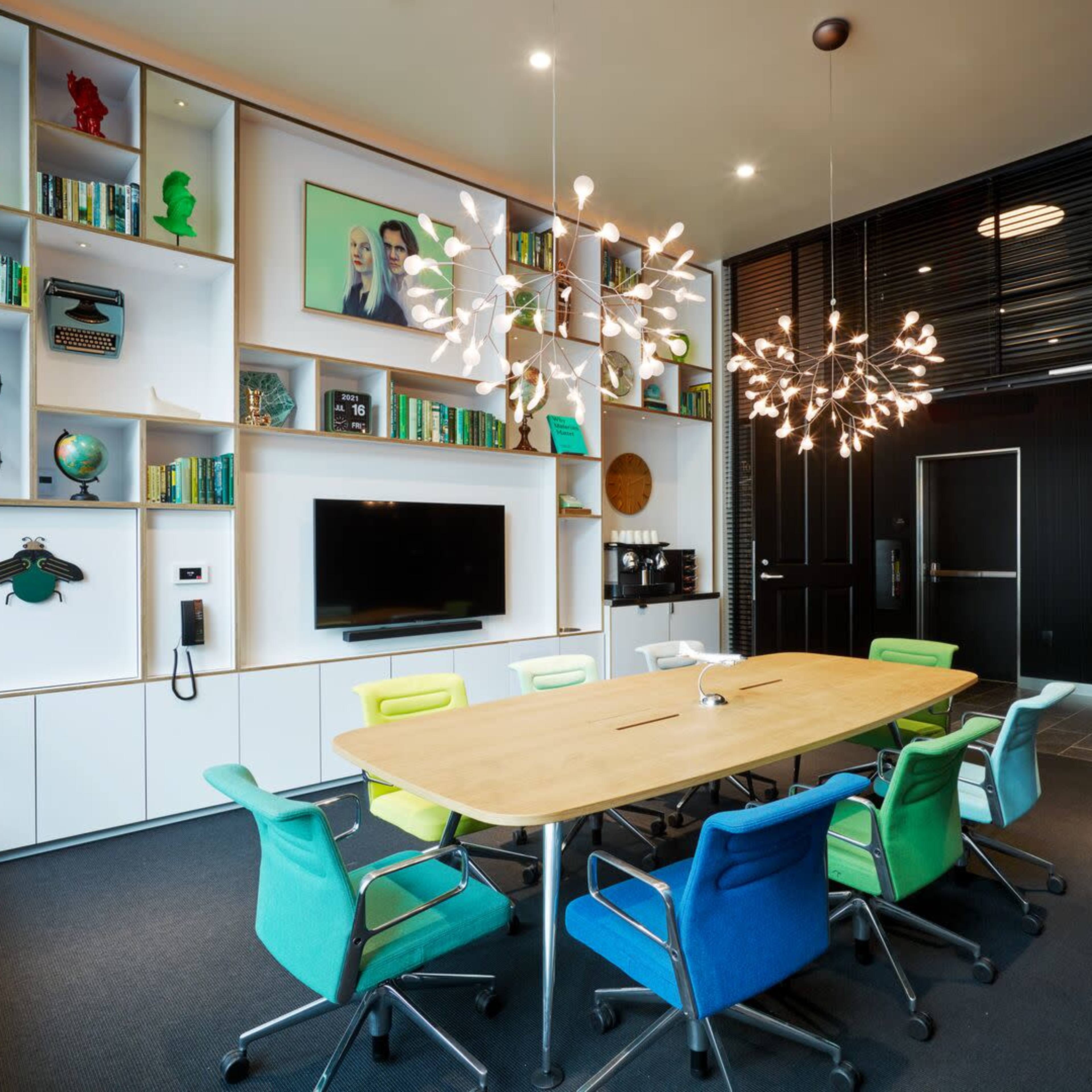 A modern meeting room features a large wooden table surrounded by colorful chairs, with a bookshelf displaying decor and books, and two distinctive light fixtures hanging from the ceiling.