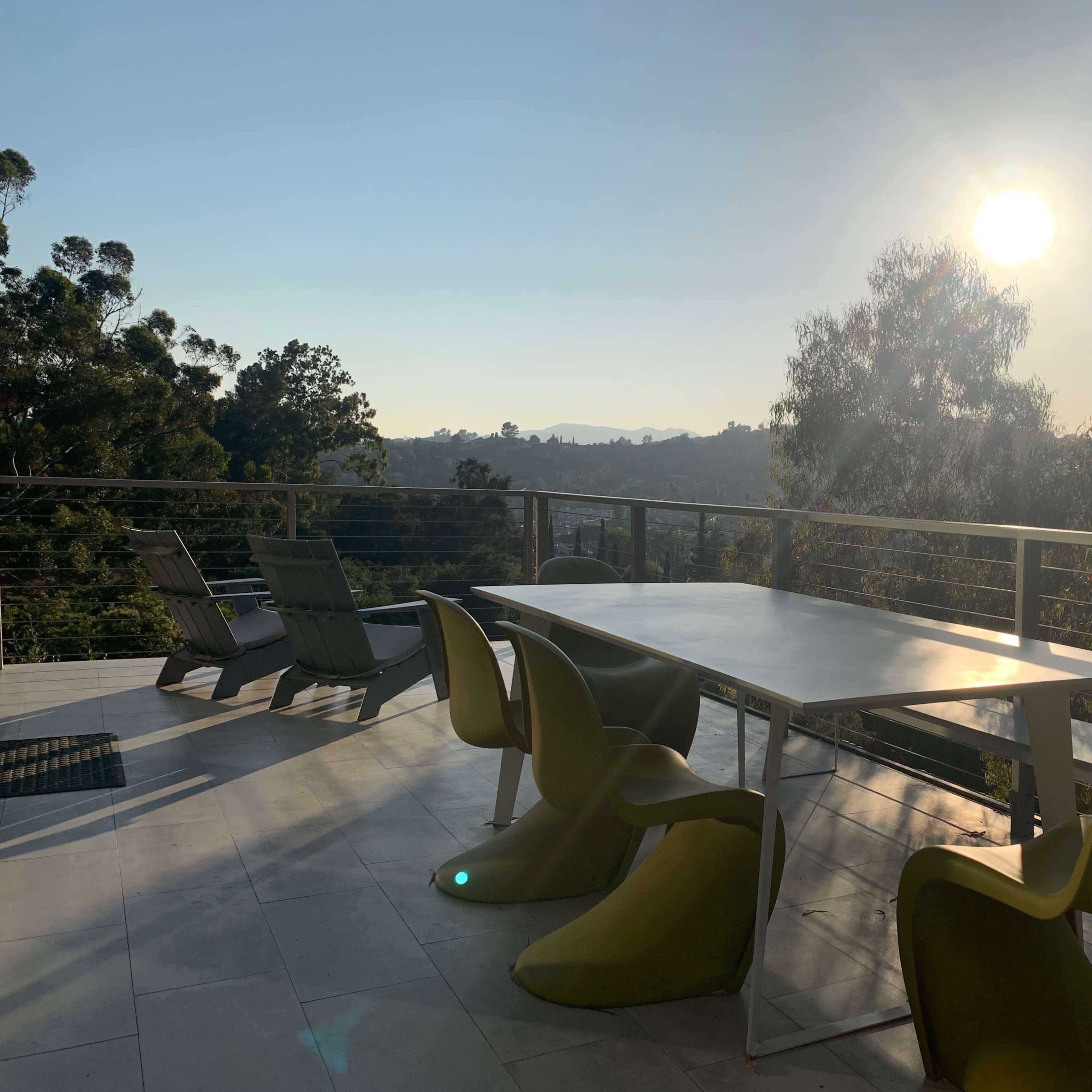 A table with colorful chairs is set on a patio overlooking a hilly landscape under a bright sun.