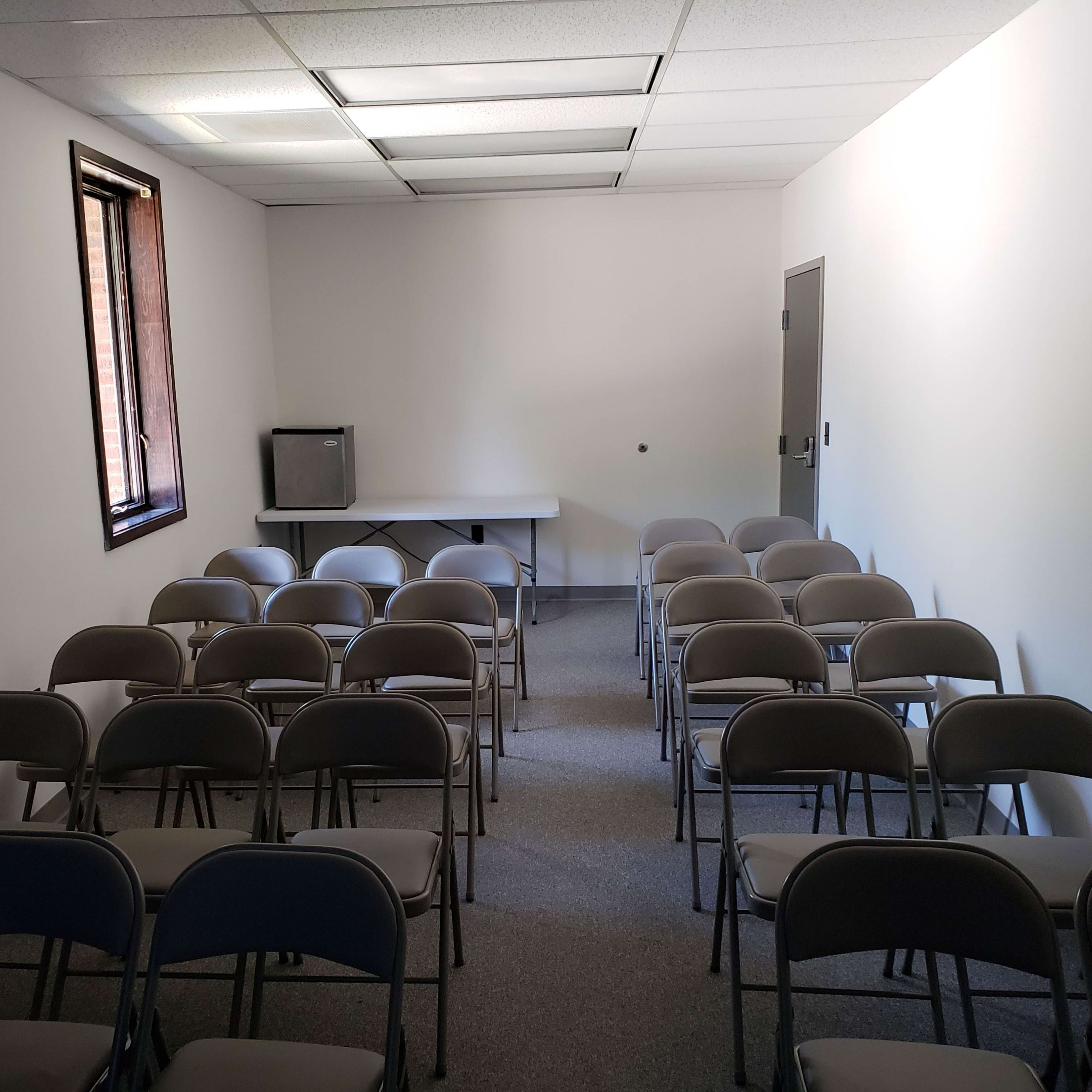 A small, empty room arranged with rows of folding chairs facing a table, with a window and a television in the corner.