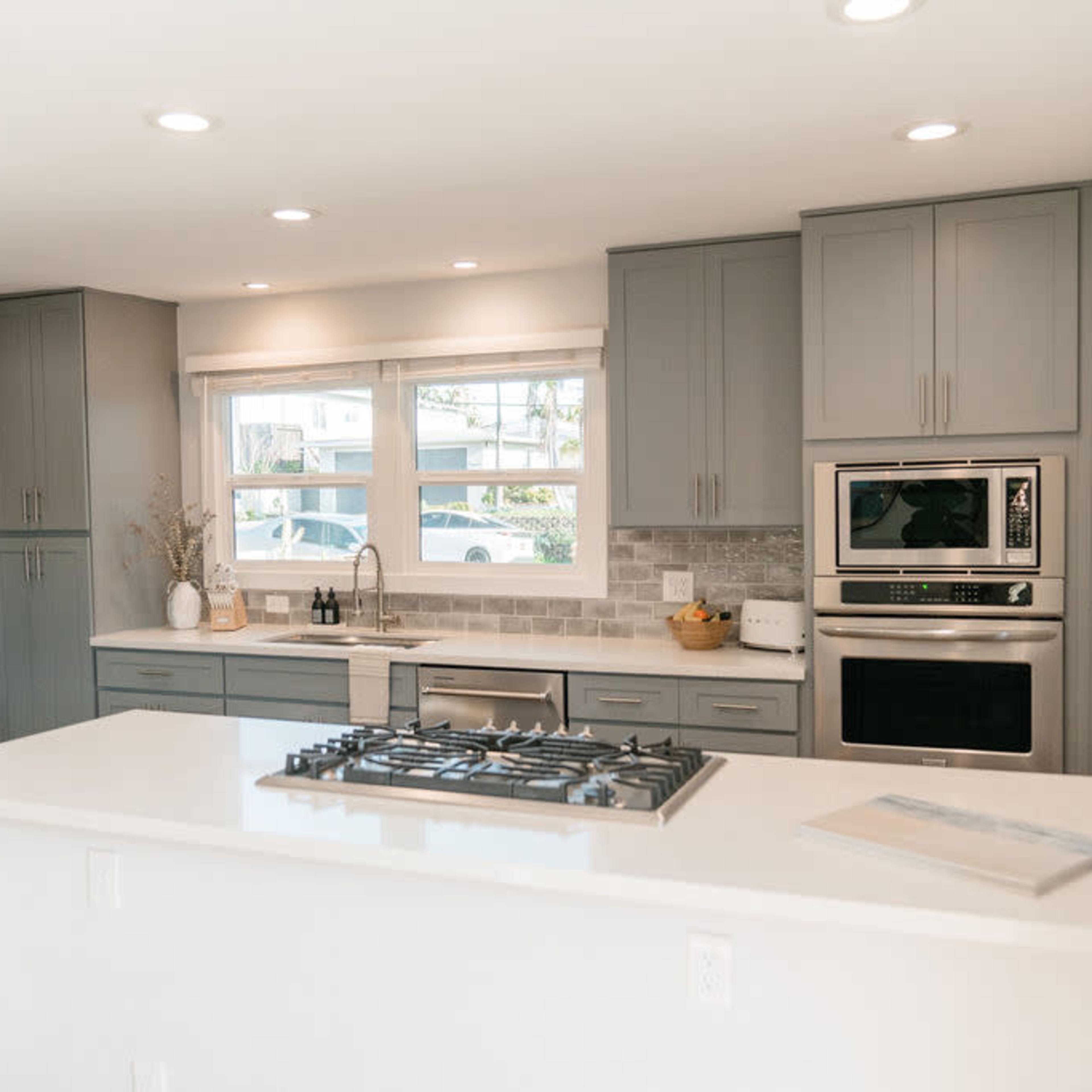 A modern kitchen with gray cabinets, a white countertop, and a gas cooktop, featuring stainless steel appliances and large windows for natural light.