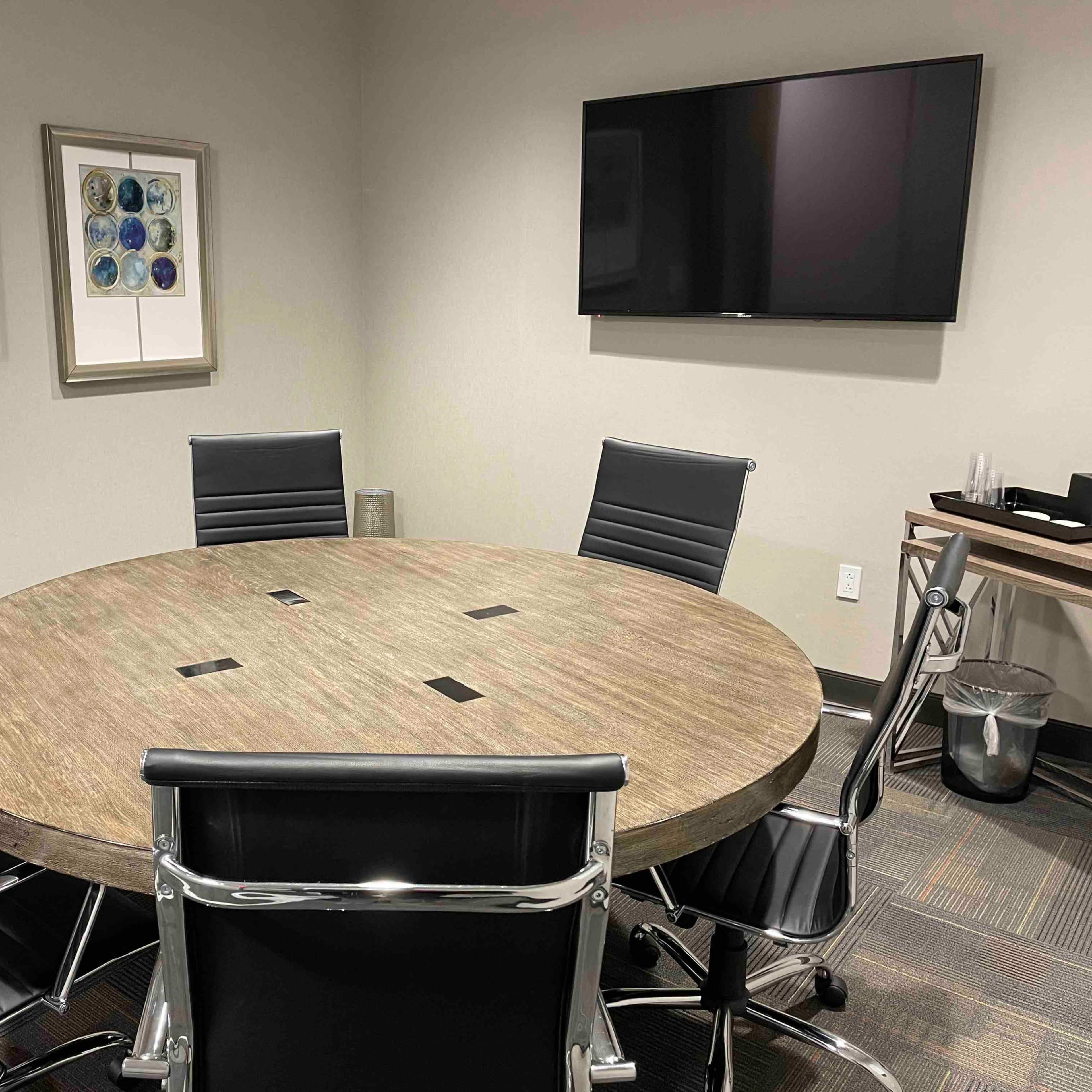 A round wooden conference table is surrounded by six black leather chairs in a meeting room with framed artwork and a wall-mounted television.