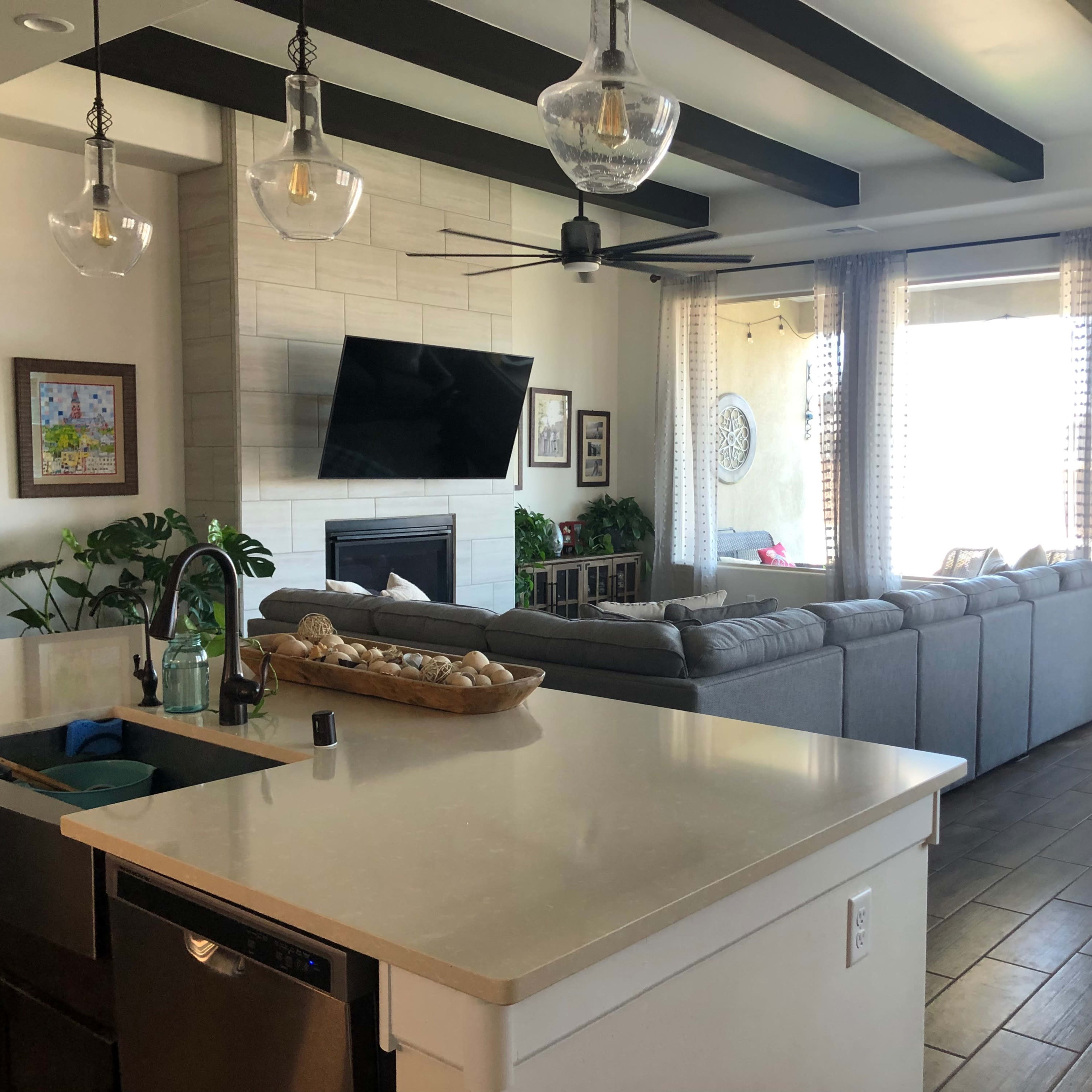 A modern living space featuring a kitchen area with a countertop, a living room with a large gray sofa, and a dining area with a wooden table, all illuminated by natural light.