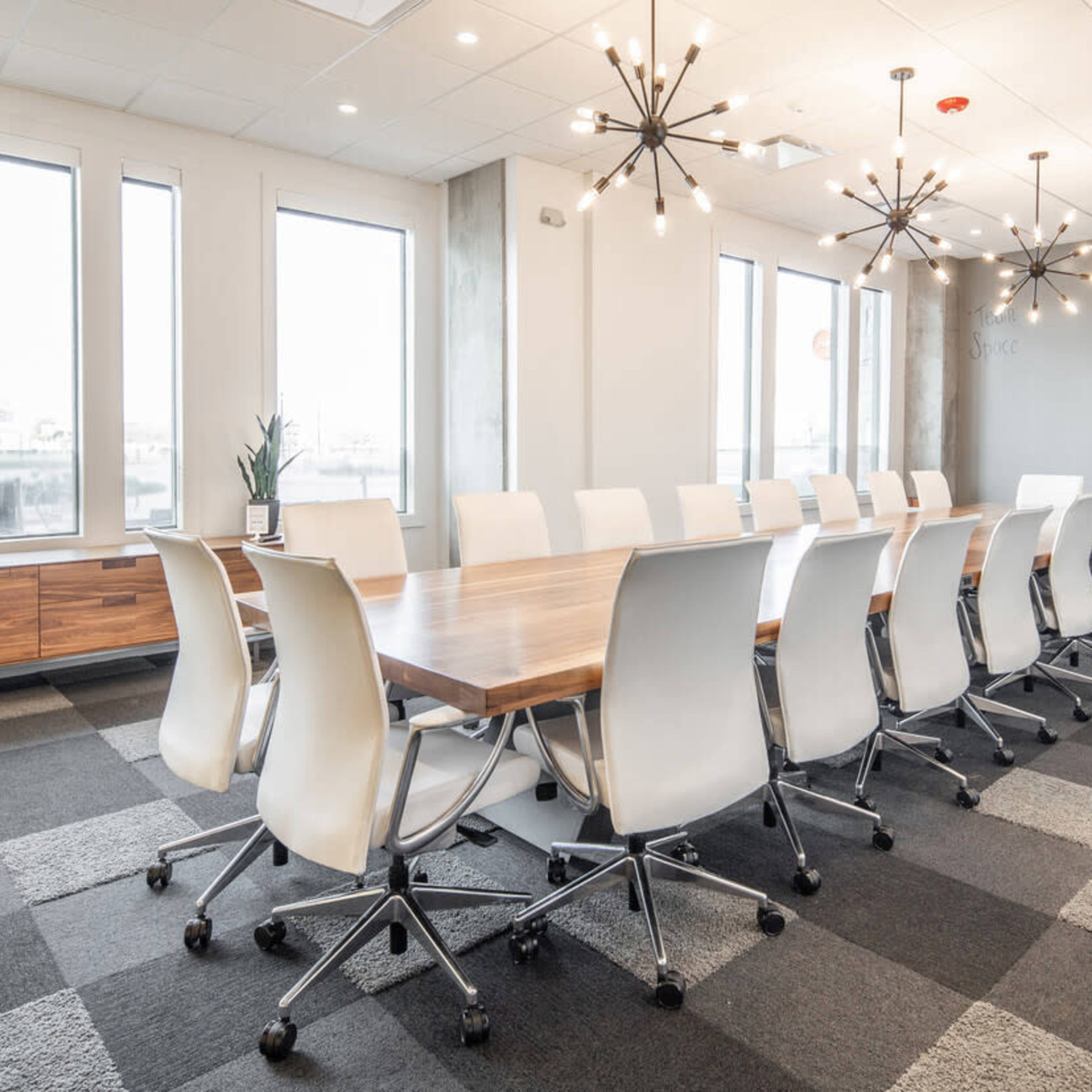 A modern conference room features a long wooden table surrounded by white swivel chairs, with large windows and contemporary light fixtures overhead.