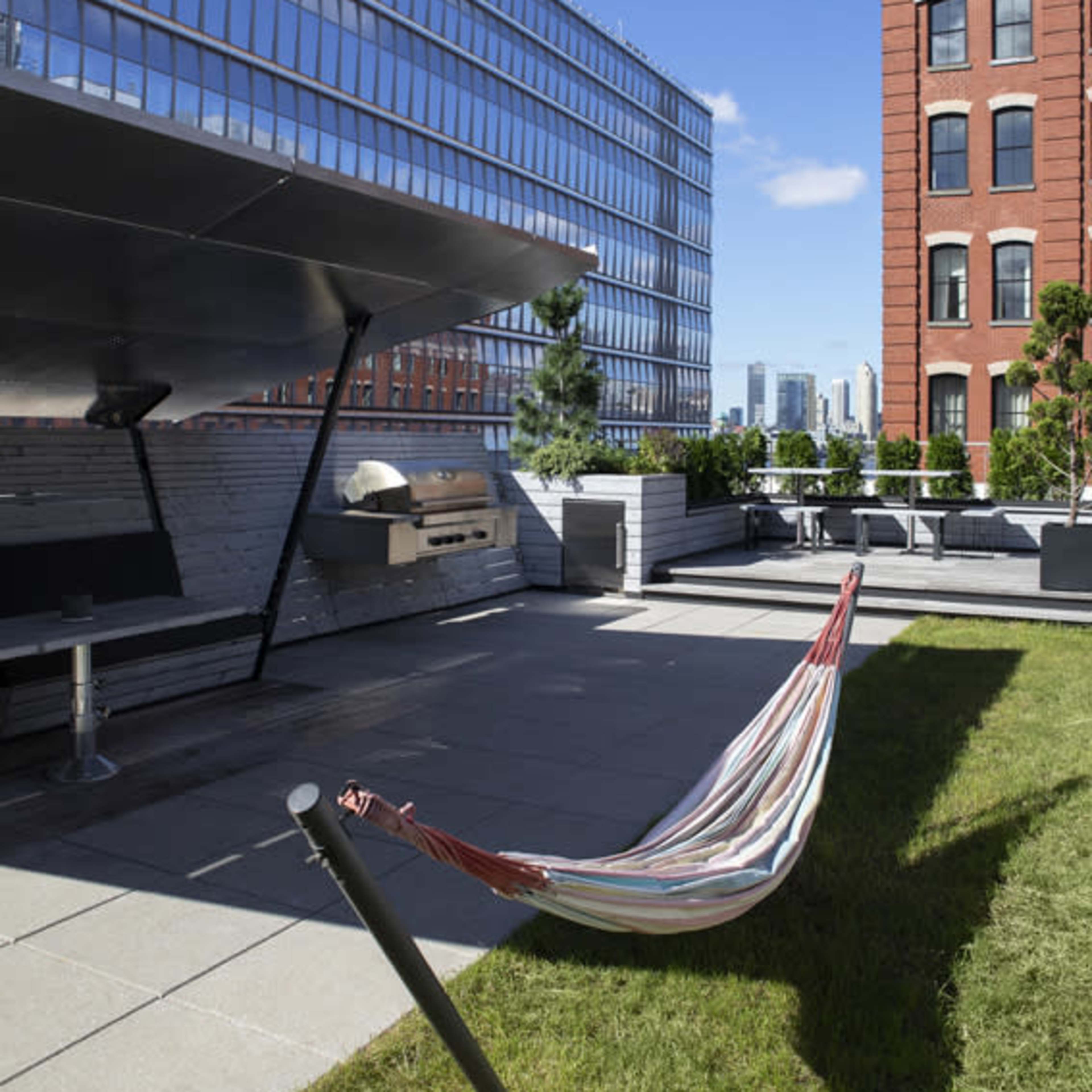A striped hammock is suspended between two supports on a rooftop terrace, surrounded by modern buildings and greenery.