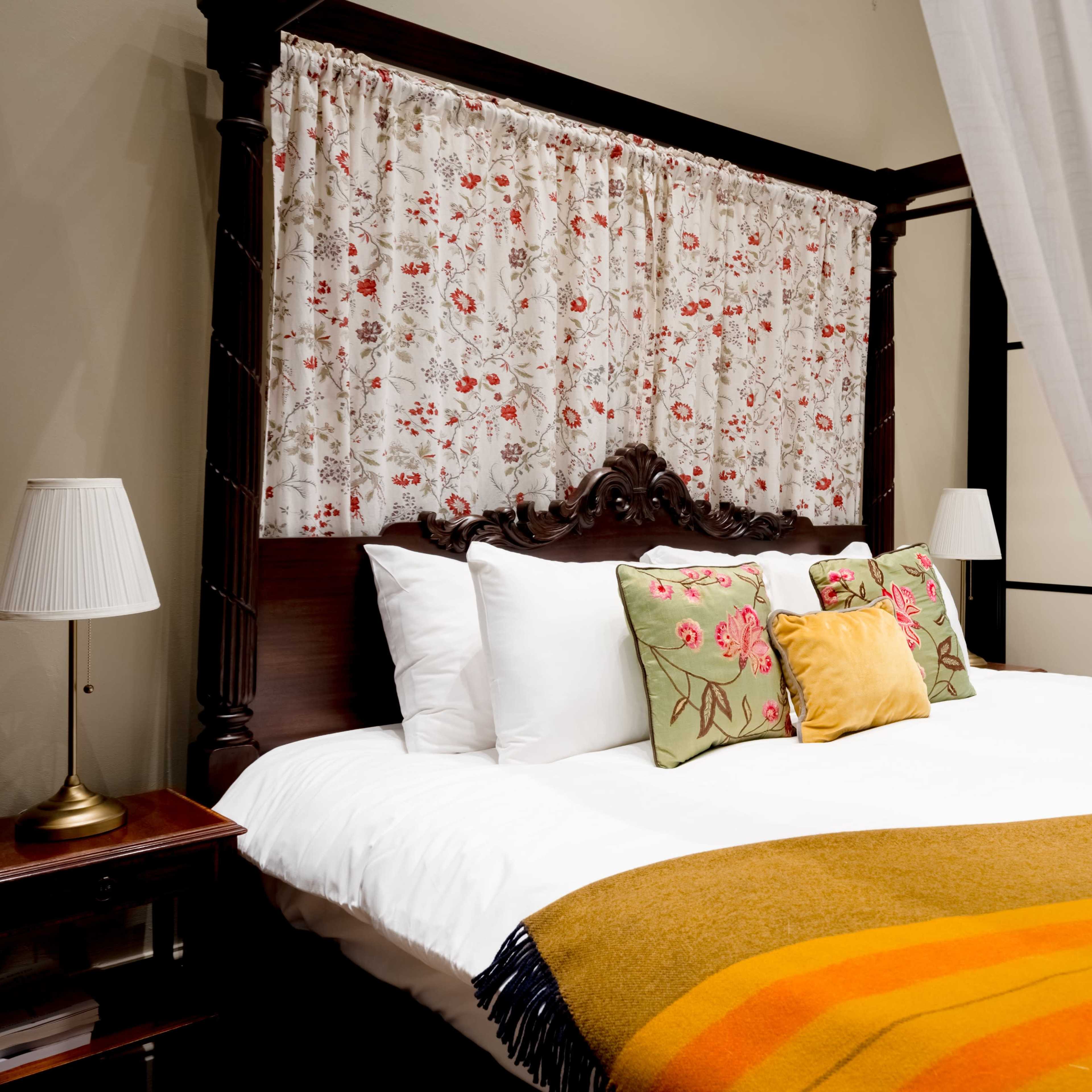 A canopy bed with a floral curtain, colorful pillows, and a blanket draped over the edge in a softly lit room.