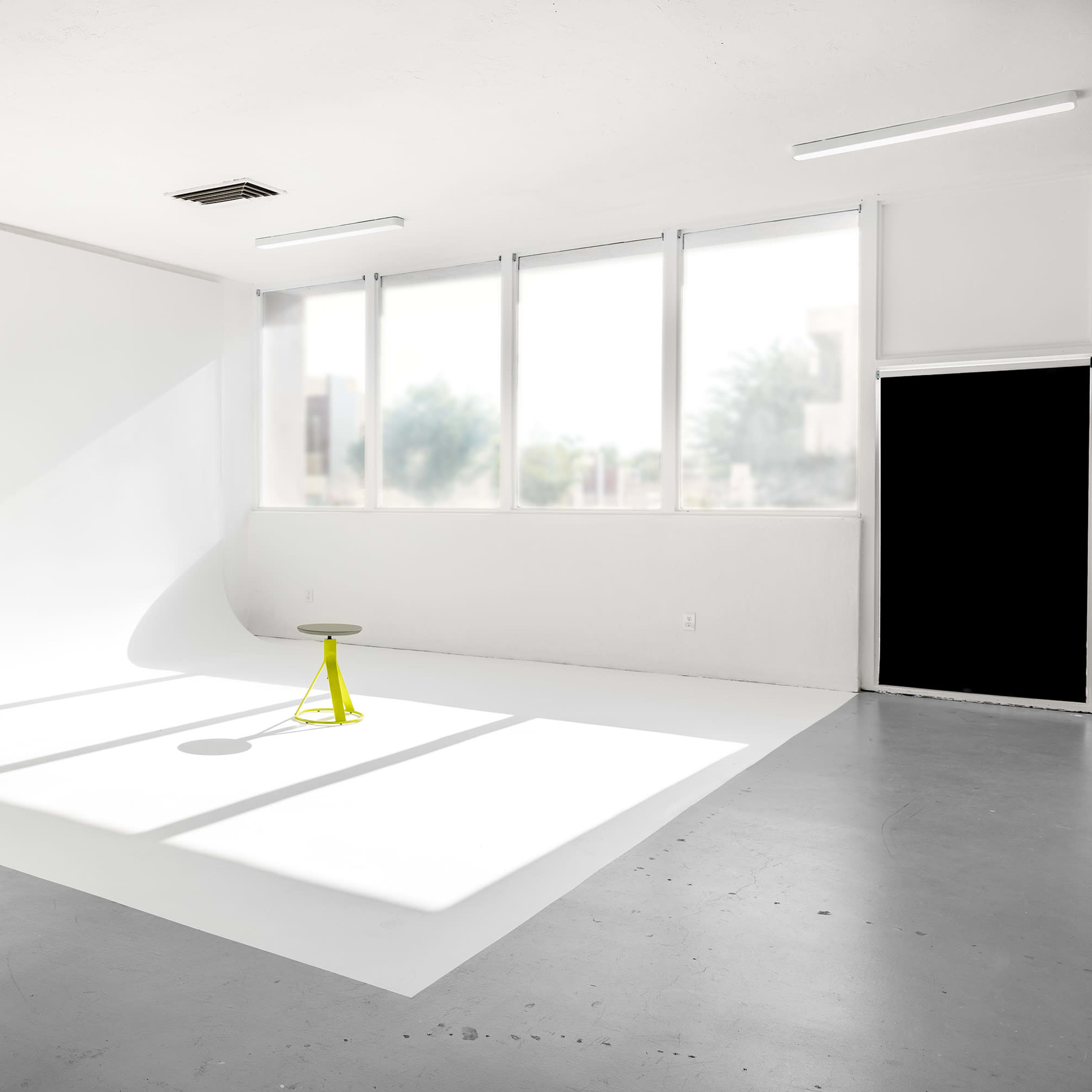 A minimalist photography studio features a yellow stool on a white backdrop with natural light streaming through large windows.