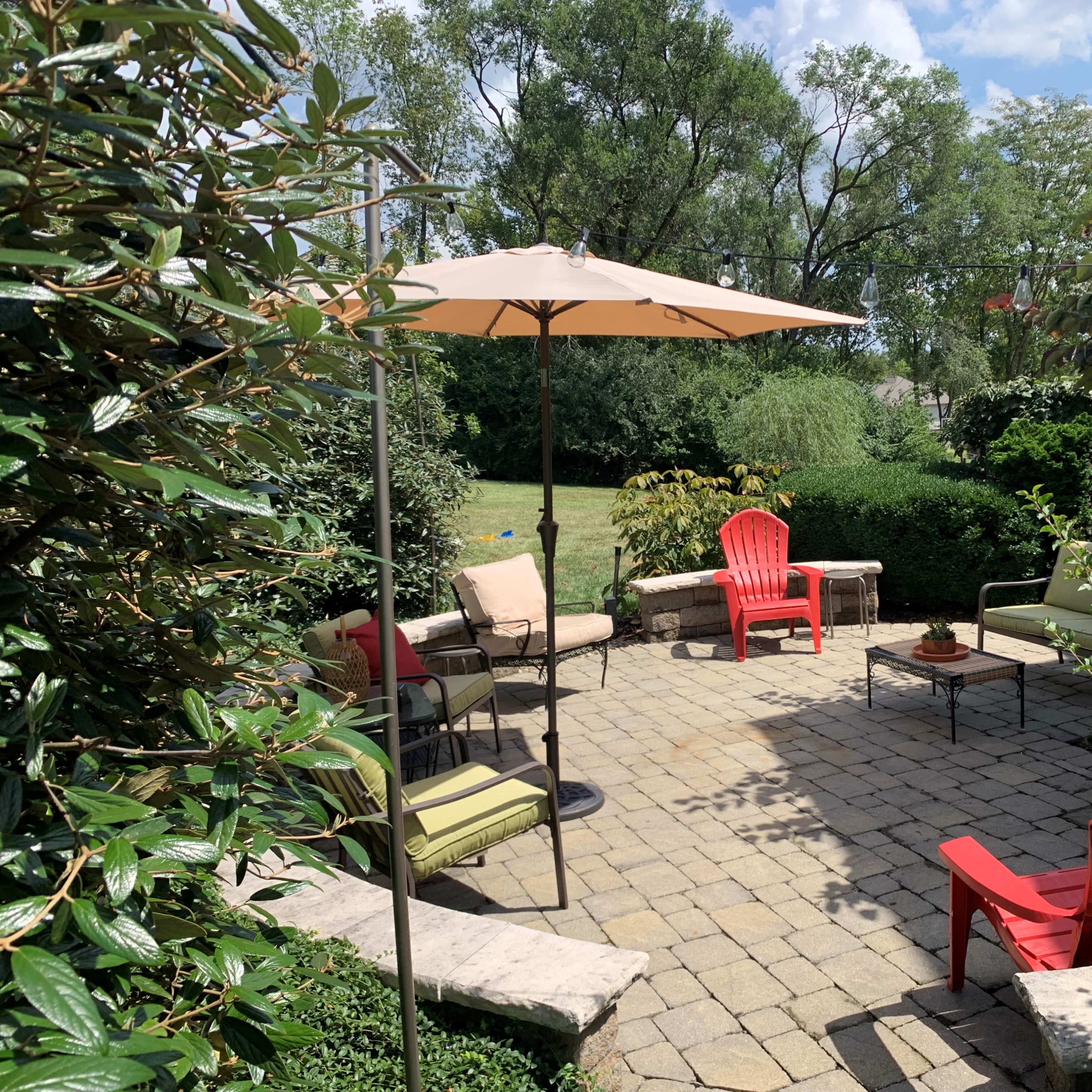 A landscaped patio with seating arrangements, an umbrella, and a stone fire pit surrounded by greenery.