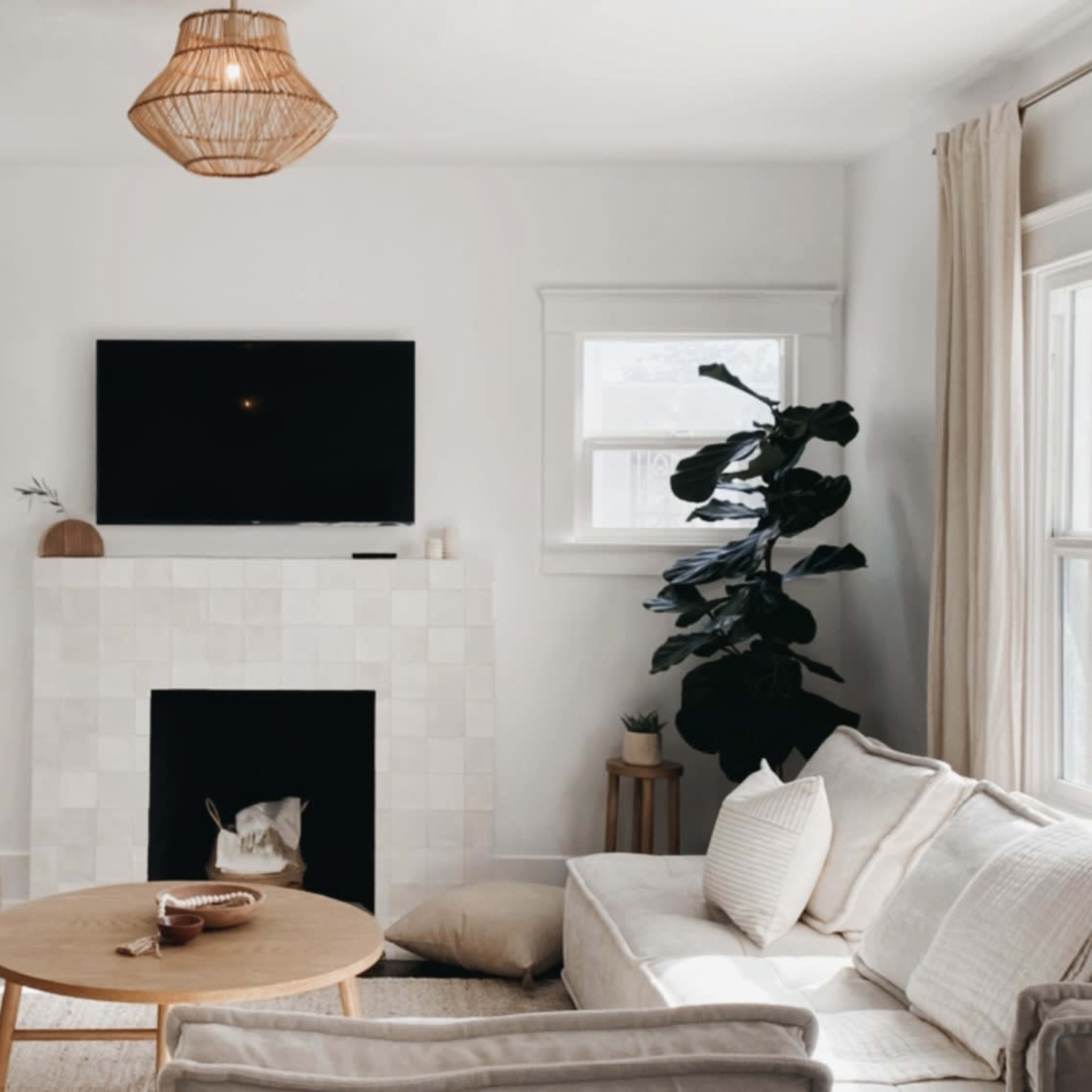 A cozy living room features a light-colored sofa and armchairs, a round wooden coffee table, and a fireplace with a TV above, surrounded by large windows and a potted plant.