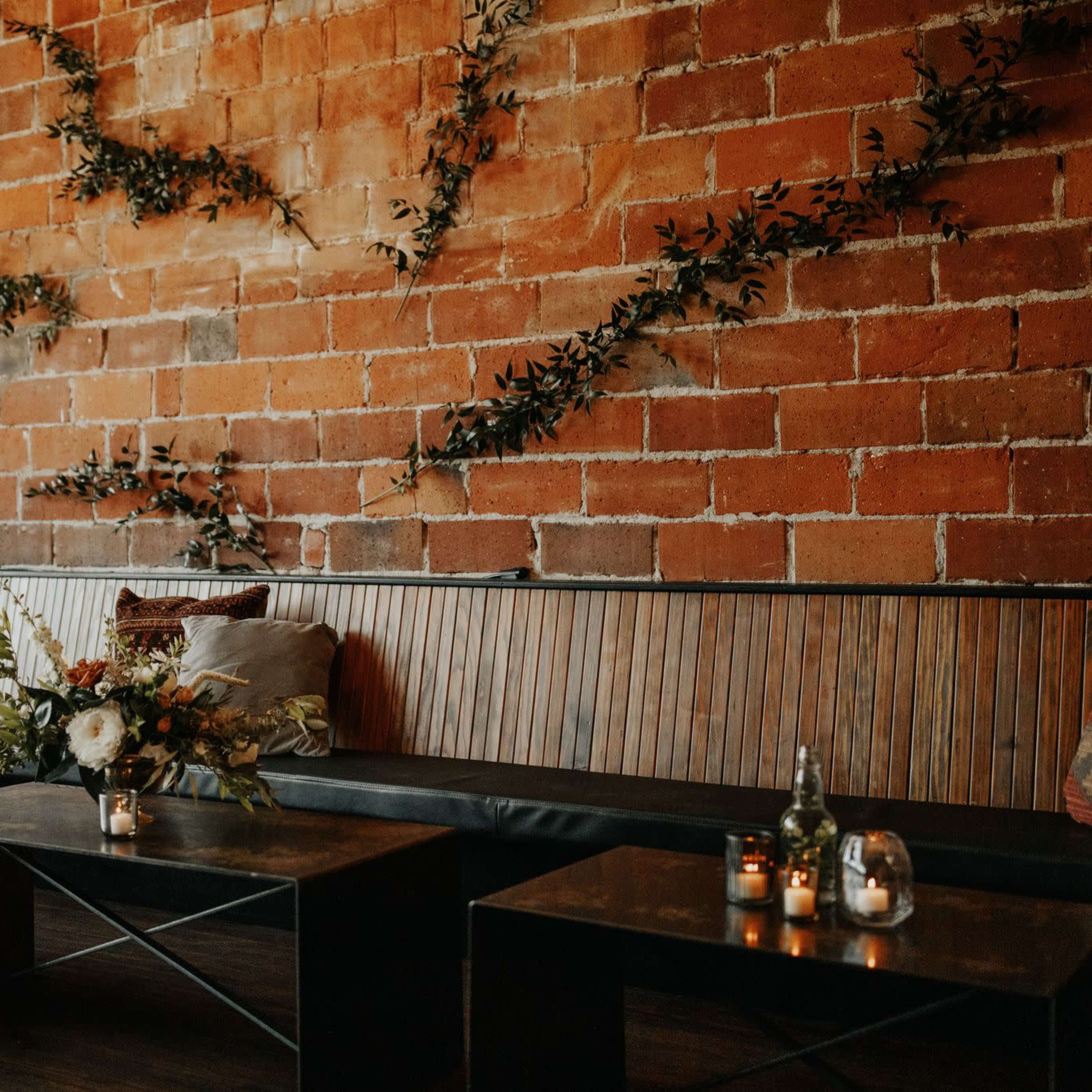 A seating area features a wooden bench against a brick wall, with decorative foliage and low, dark tables arranged in front.