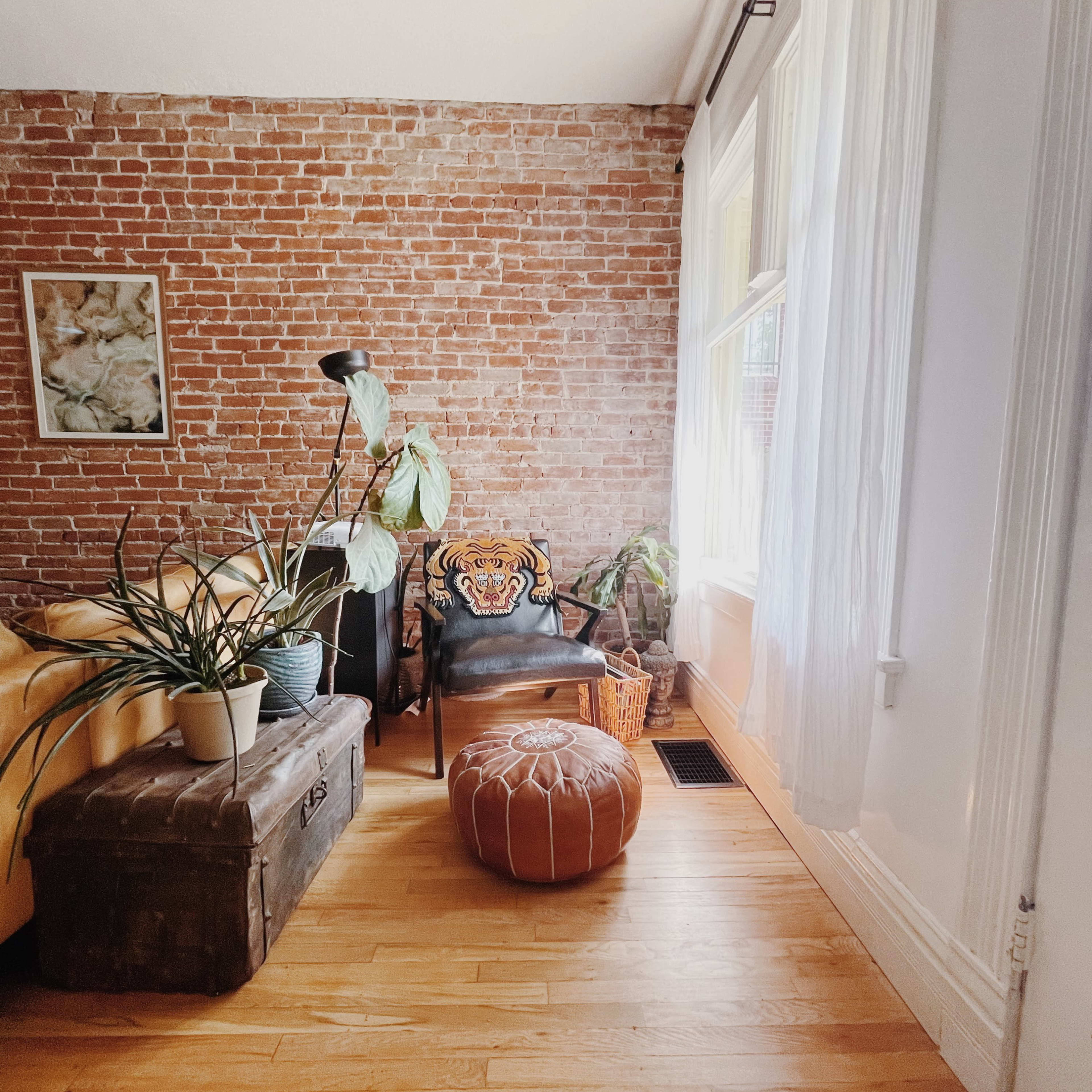 A cozy living room with brick walls, a yellow sofa, a wooden trunk, a plant, and a patterned pouf.
