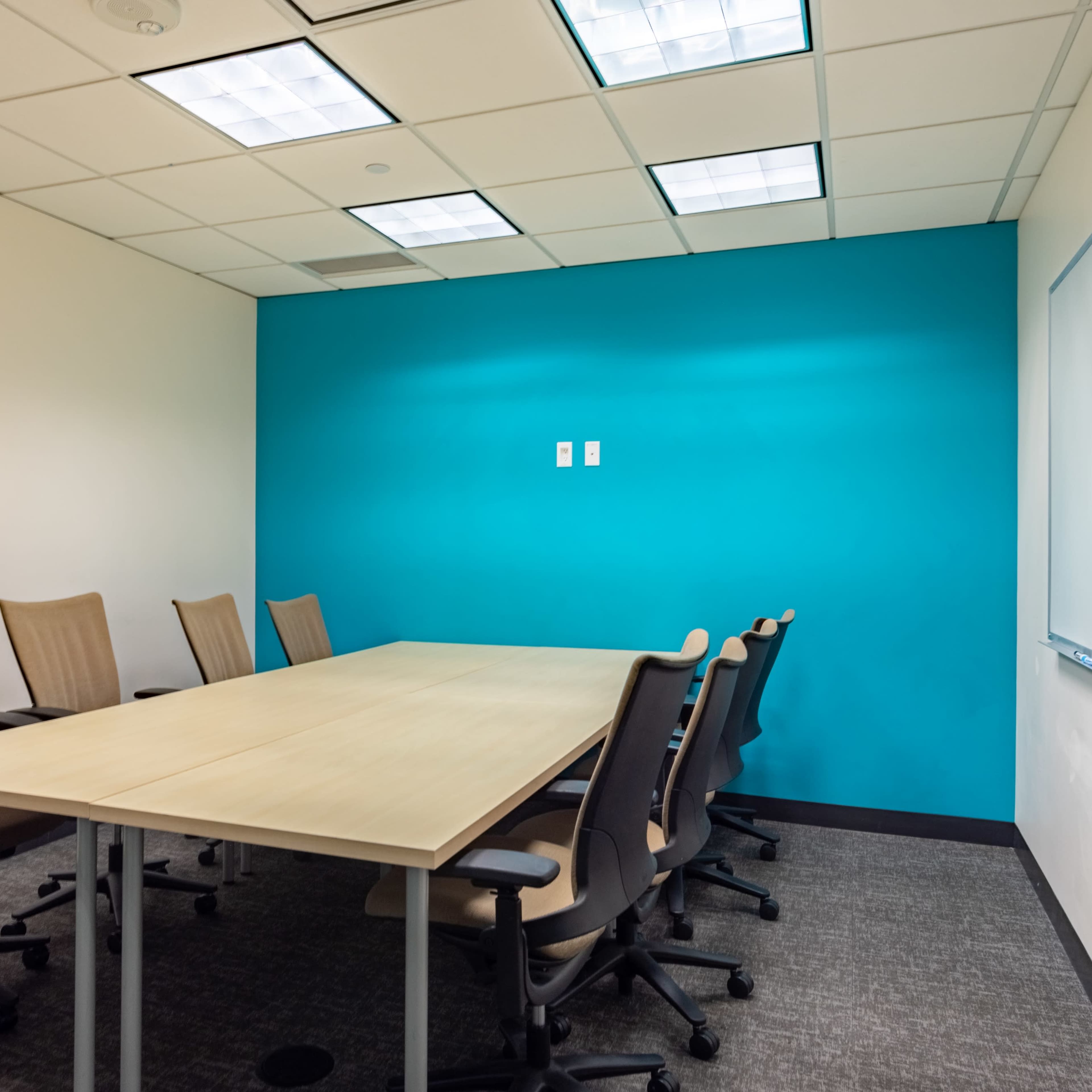 A conference room features a wooden table surrounded by eight beige chairs, with a teal accent wall and a whiteboard.