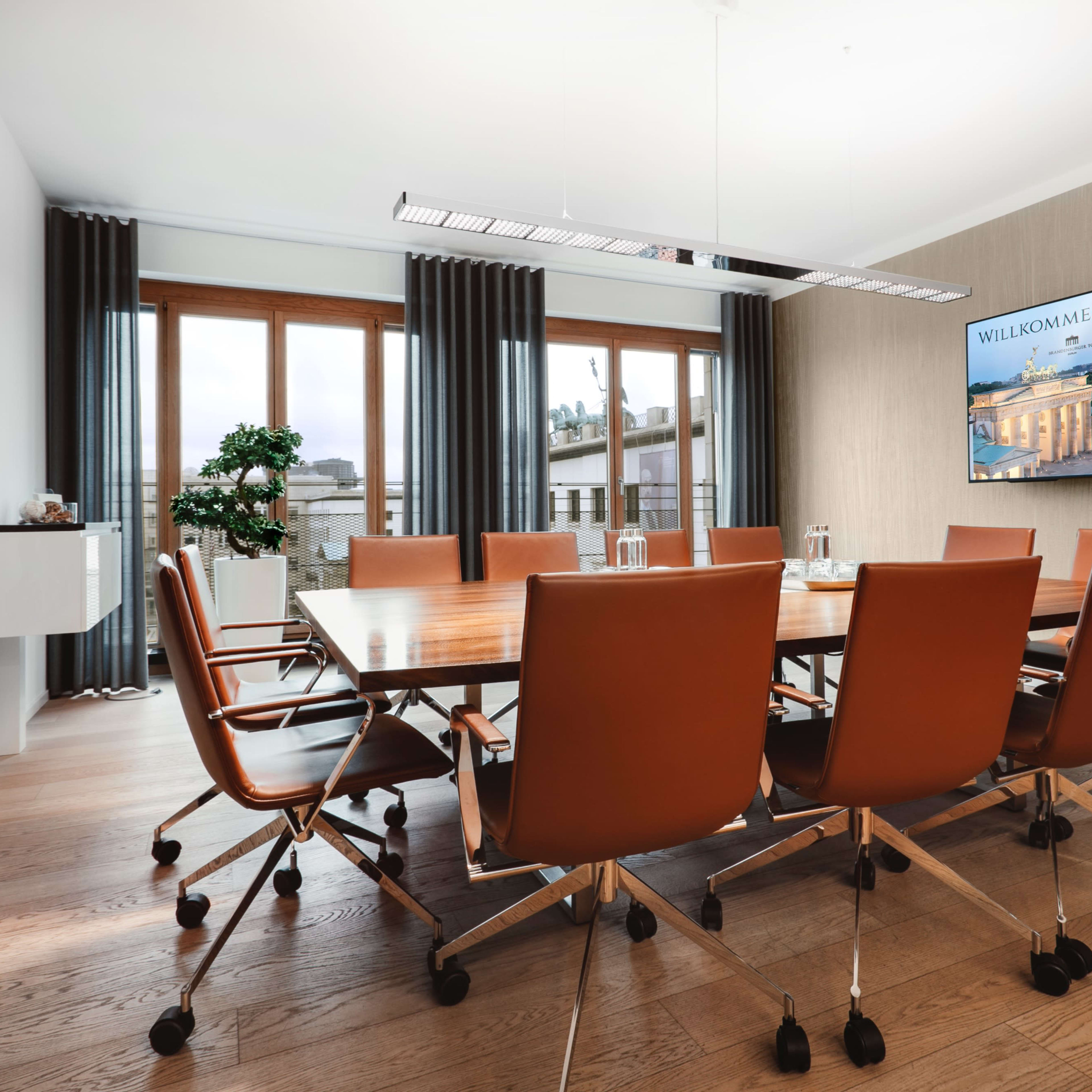 A spacious conference room features a large wooden table surrounded by orange leather chairs, with a large screen displaying a welcome message in Berlin and windows providing natural light.