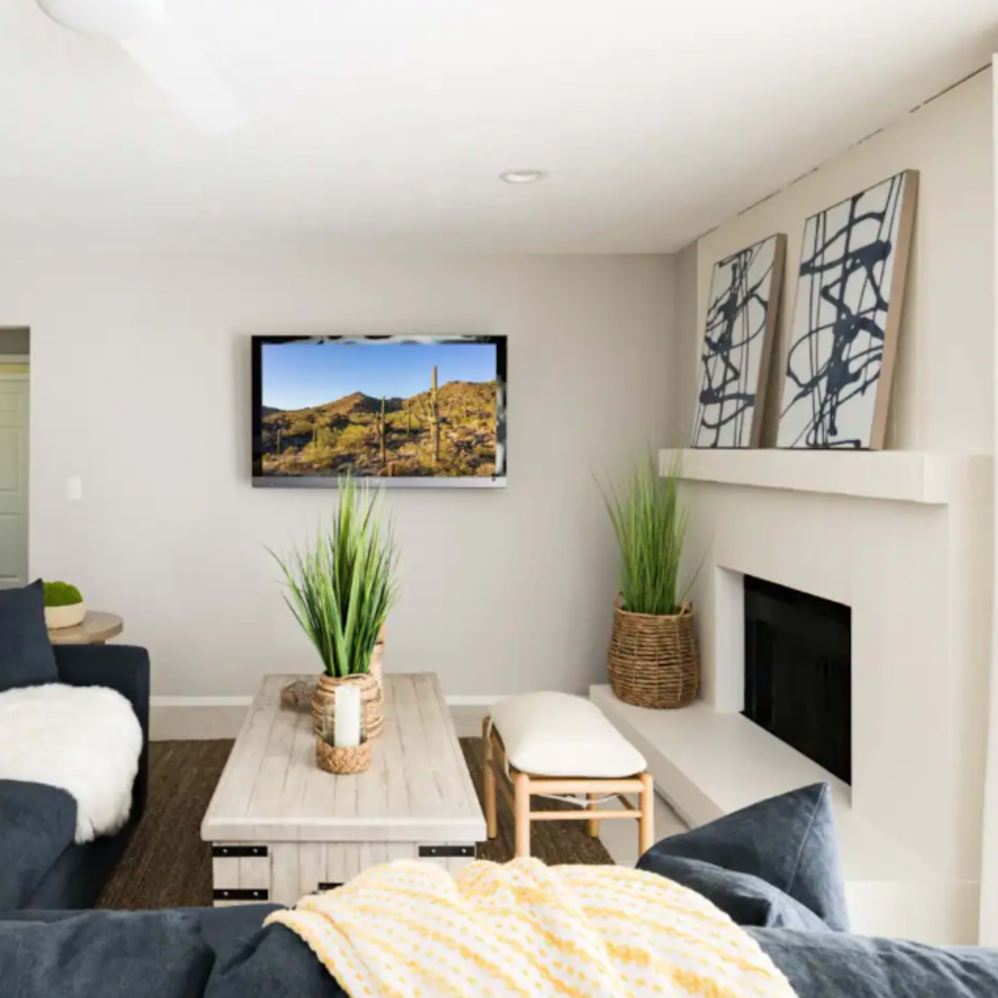 A modern living room features a dark sofa, a wooden coffee table, potted plants, and a wall-mounted television displaying a desert landscape.