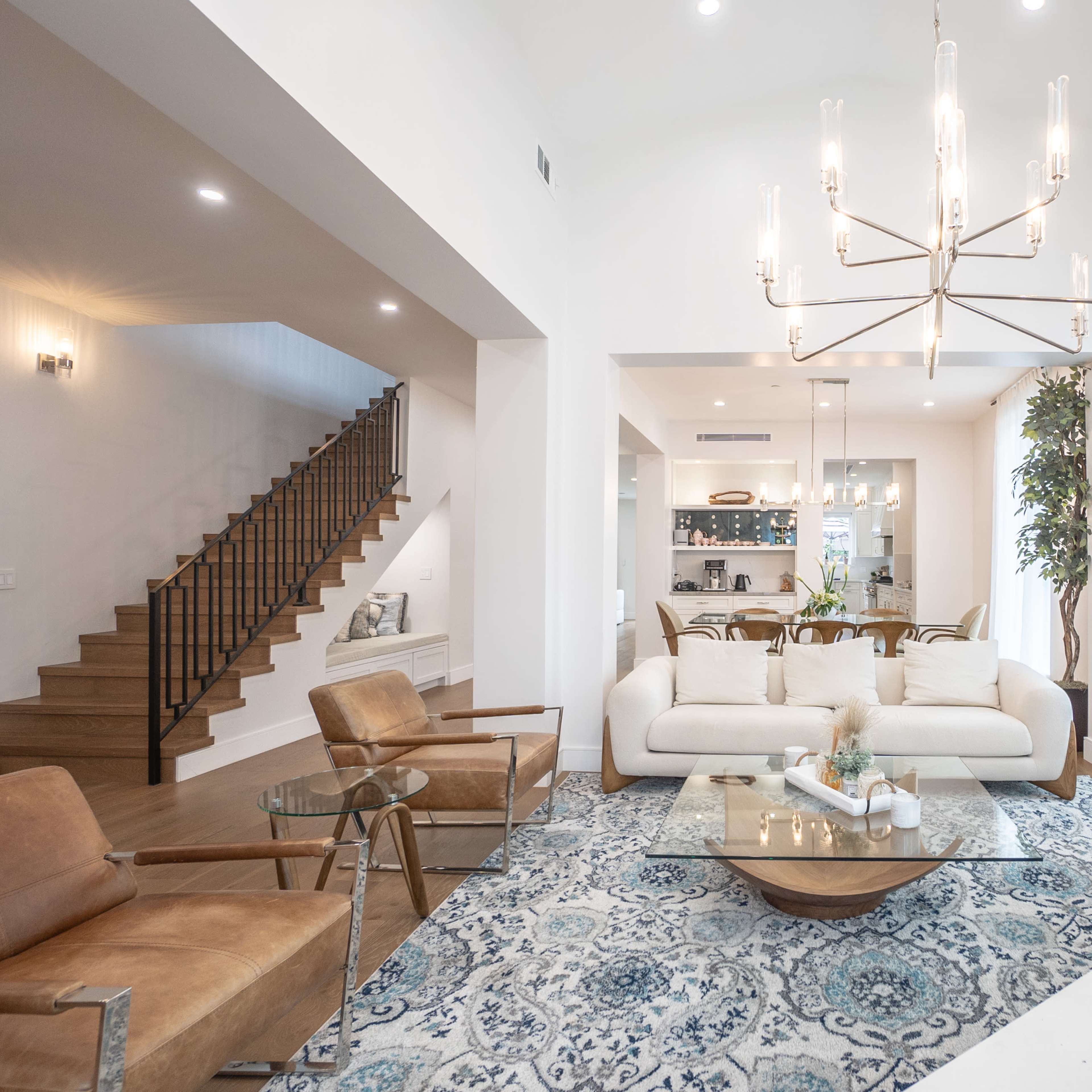 A spacious living room features a light-colored sofa, two armchairs, a glass coffee table, and a chandelier, with a staircase visible in the background.