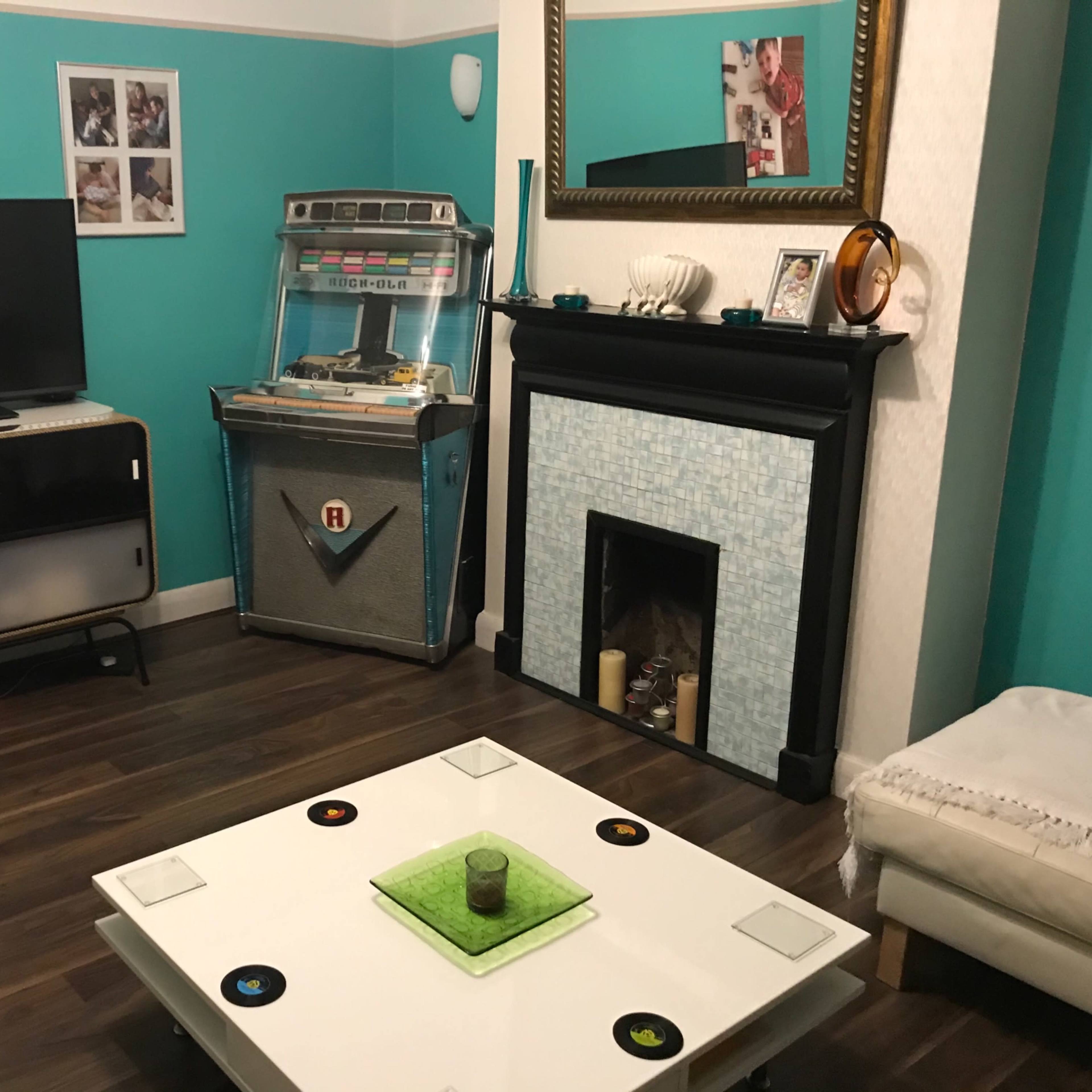 A living room with a television, a jukebox, and a coffee table, all set against teal walls and wooden flooring.