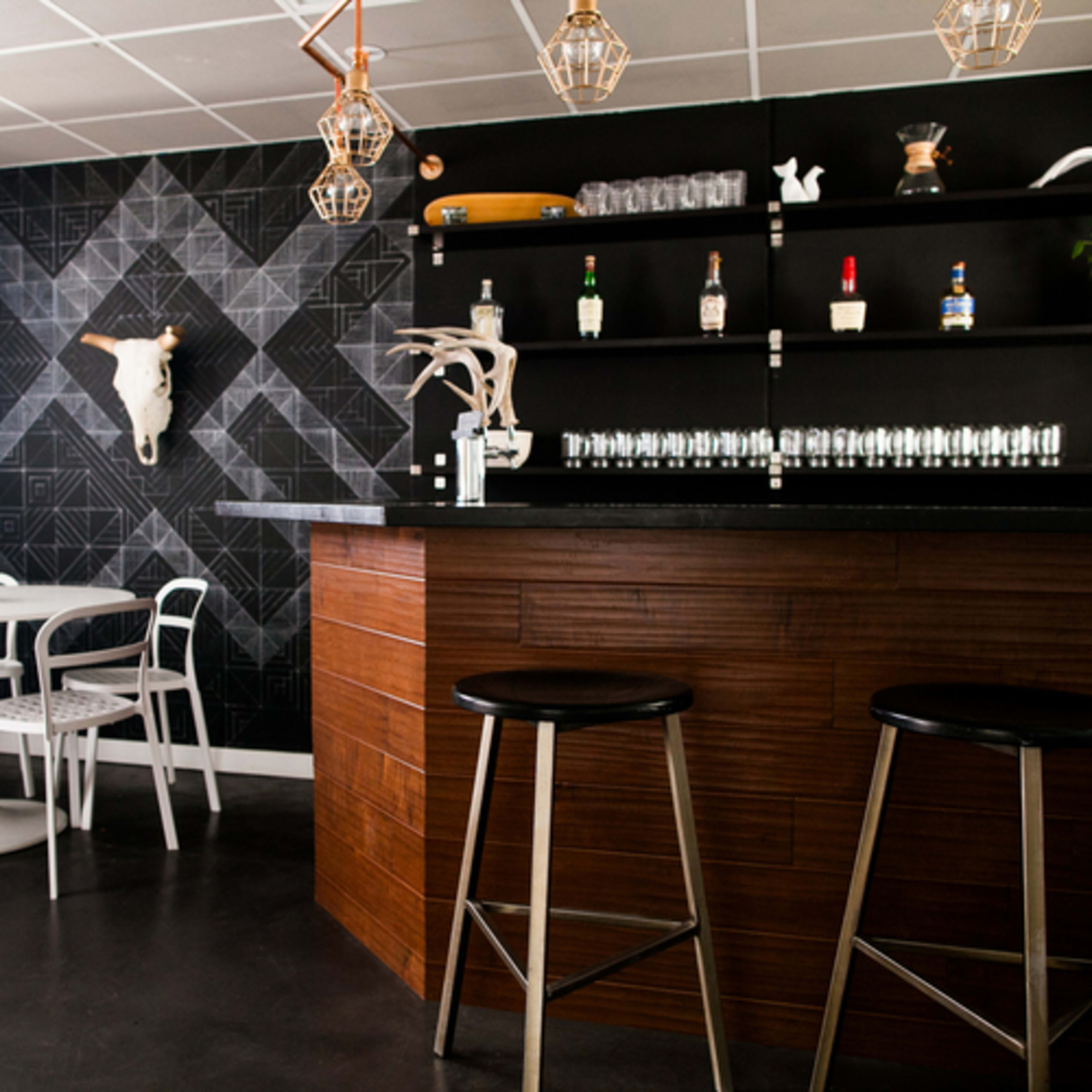 A modern bar area with a dark wooden counter, a minimalist round table with white chairs, and a geometric patterned wall adorned with a decorative skull.