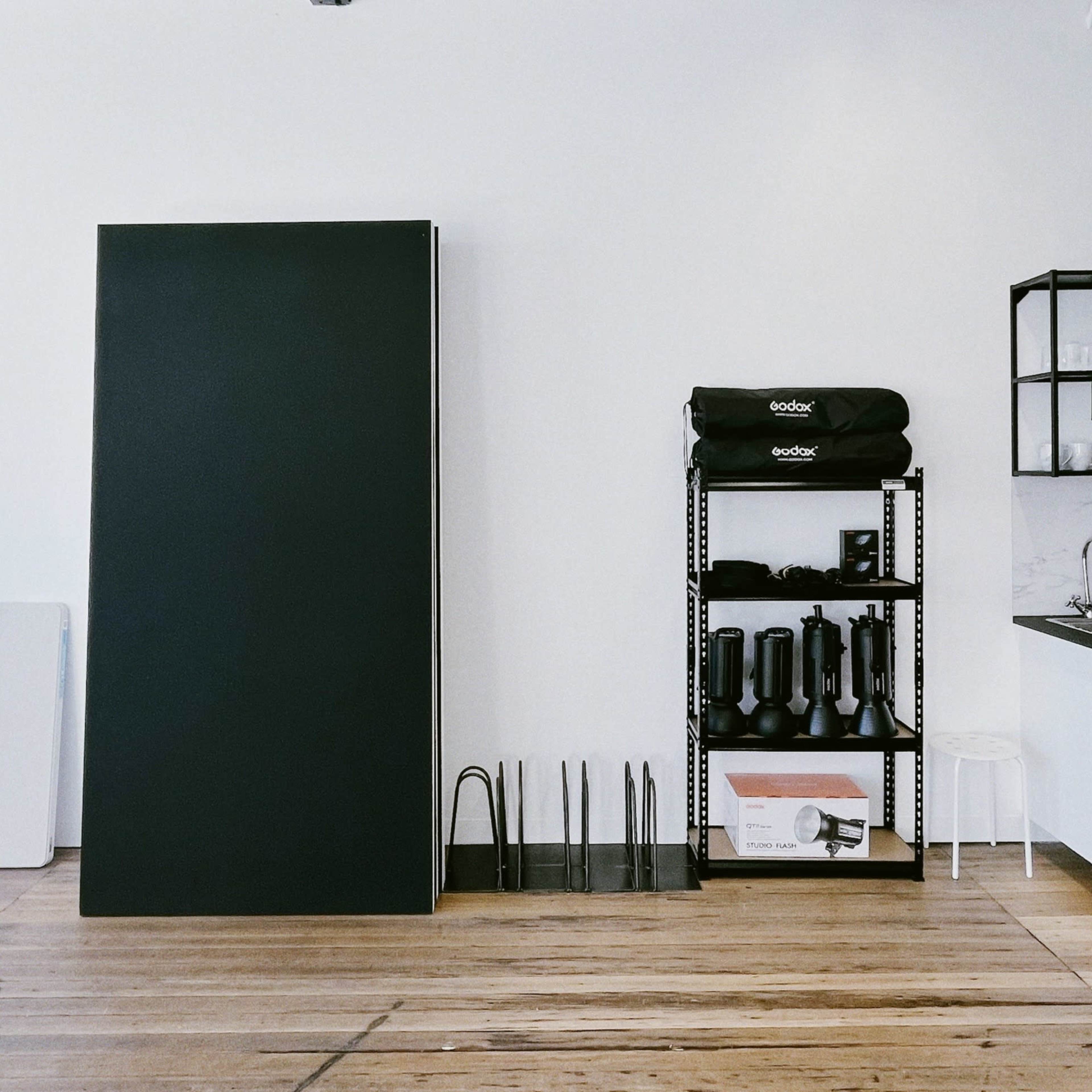 A minimalist interior space with a large black board leaning against a white wall, accompanied by a shelving unit holding various kitchen and photography equipment.