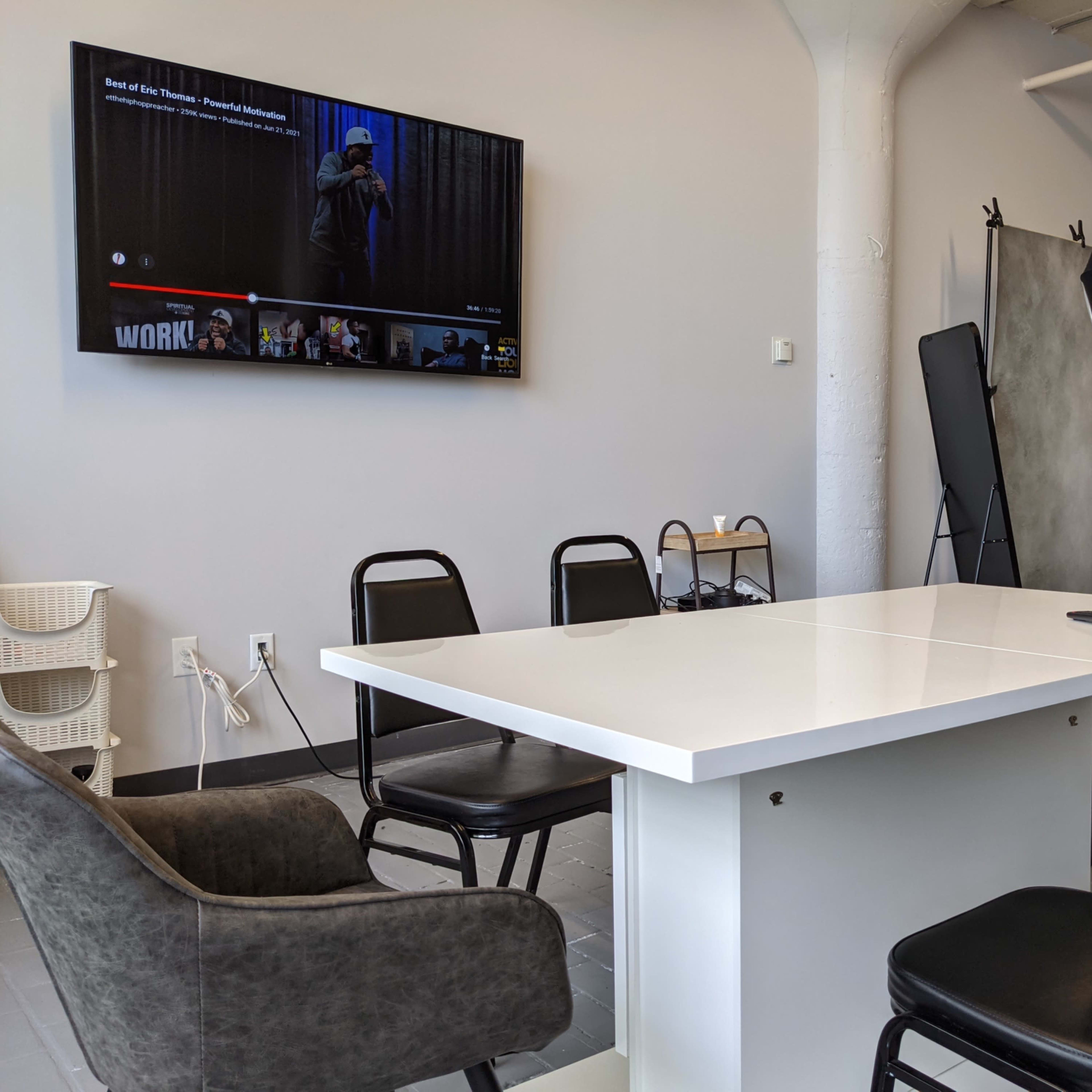 A modern conference room features a white table surrounded by six chairs, with a television mounted on the wall displaying a paused video, and photography equipment in the background.