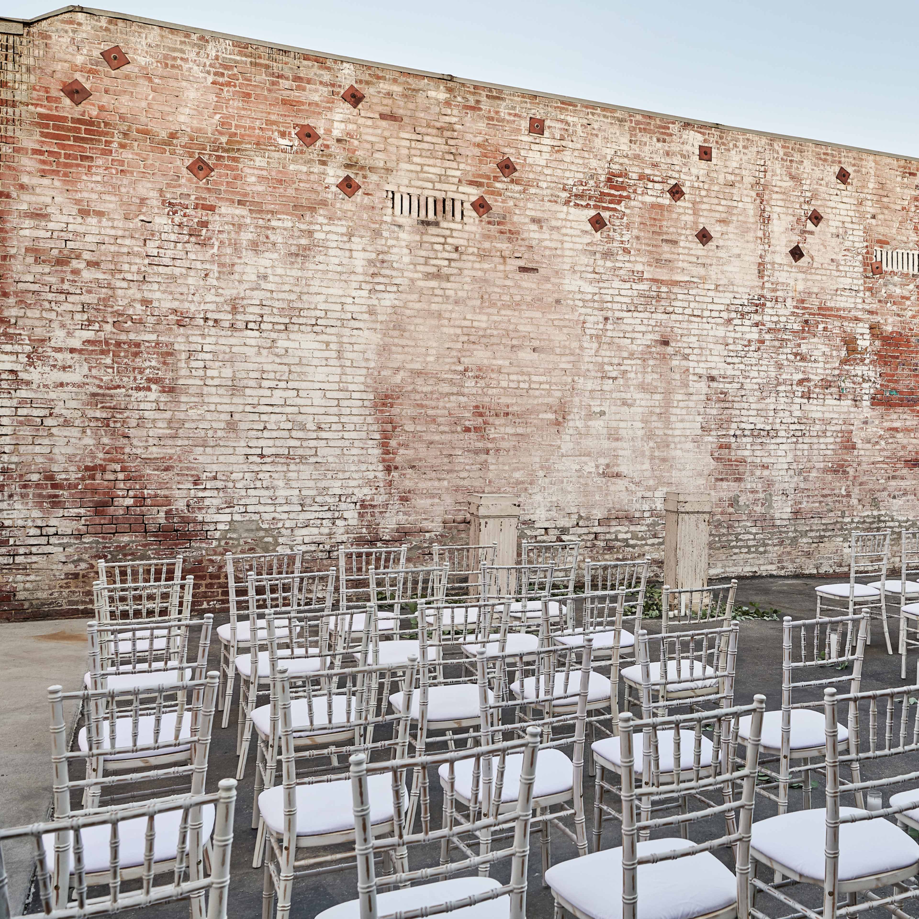 A set of white chairs arranged in rows in front of a weathered brick wall in an outdoor setting.