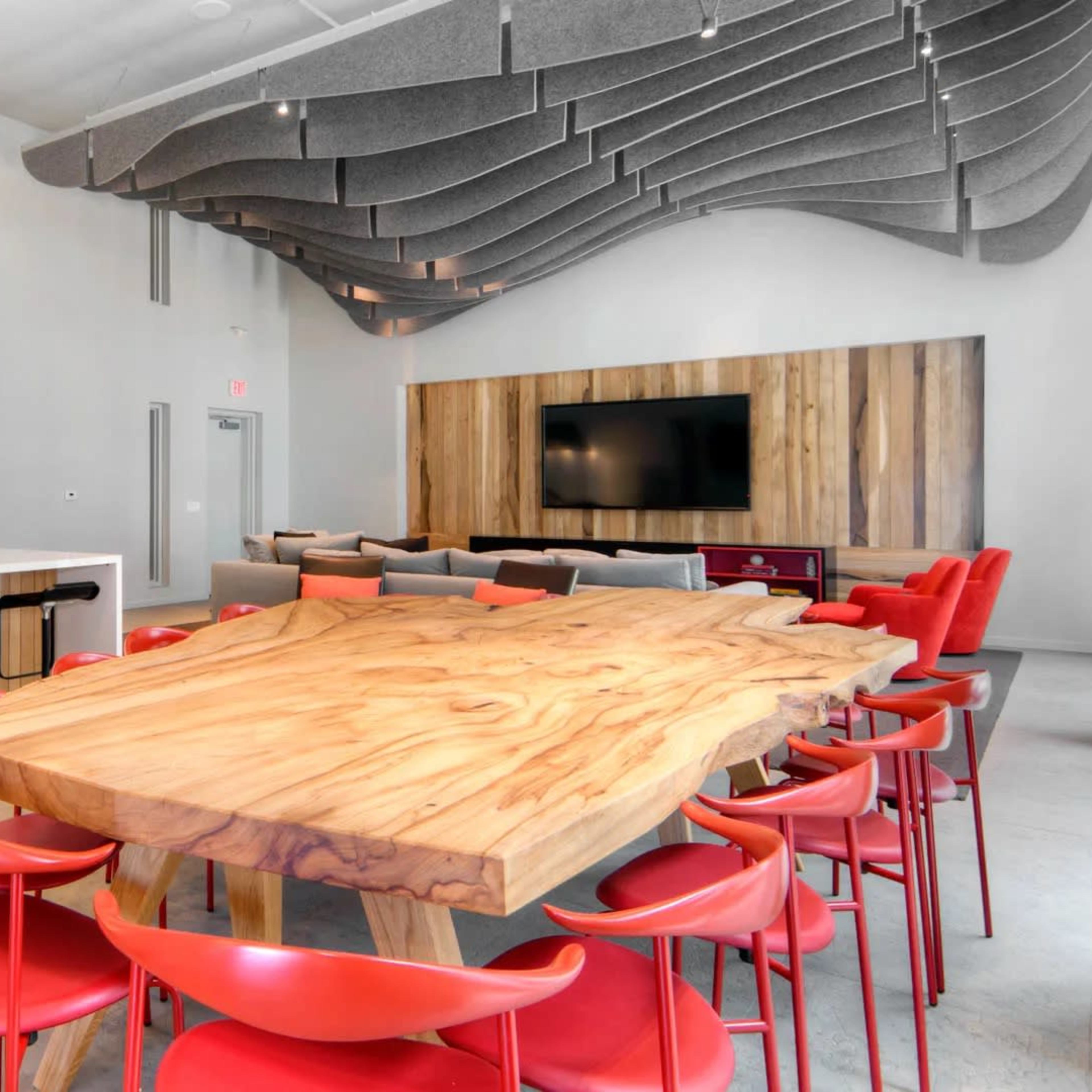 A modern dining area with a large wooden table surrounded by red chairs, a kitchen in the background, and acoustic panels on the ceiling.