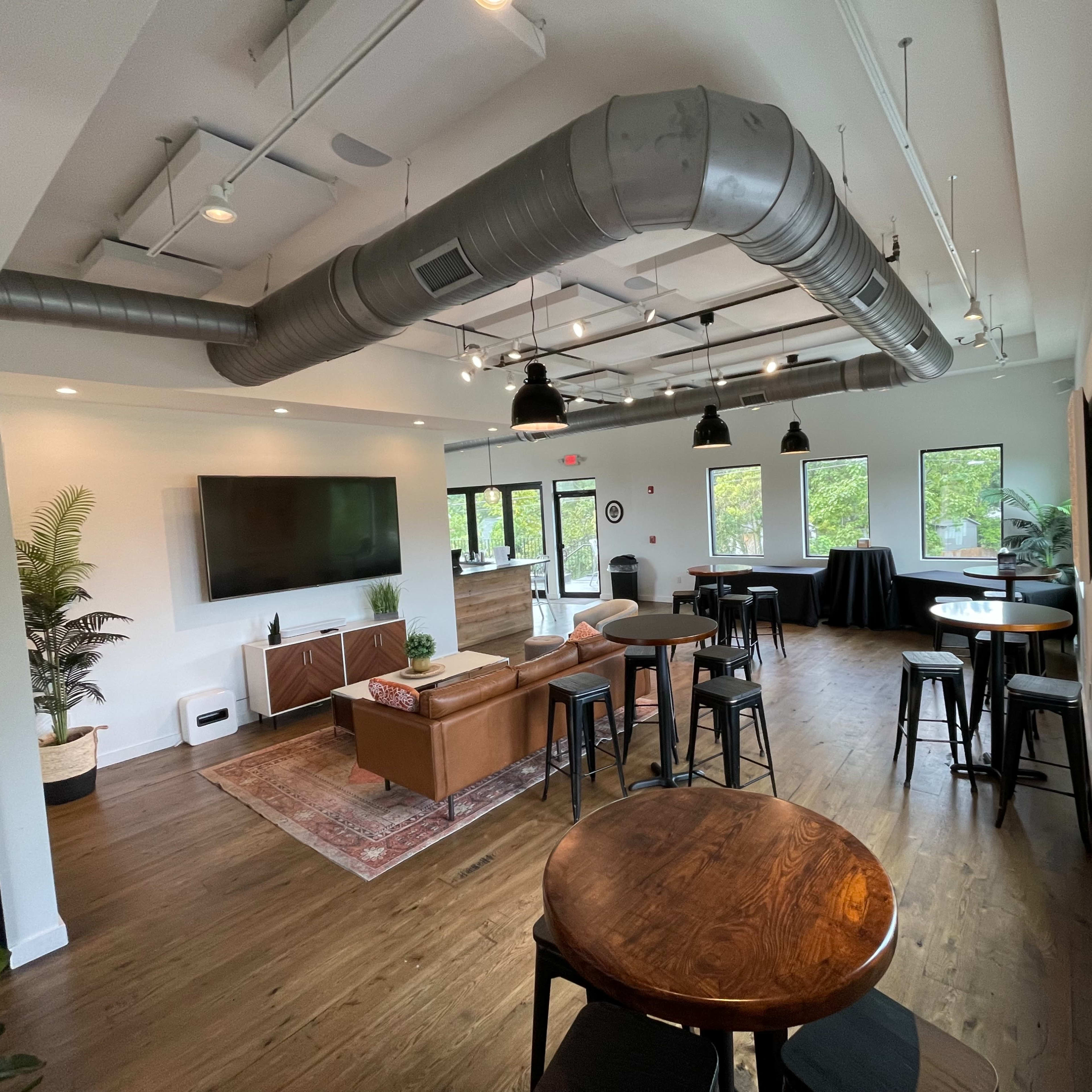 A modern lounge area with wooden floors, several tables, black chairs, a large television on the wall, and a sofa.