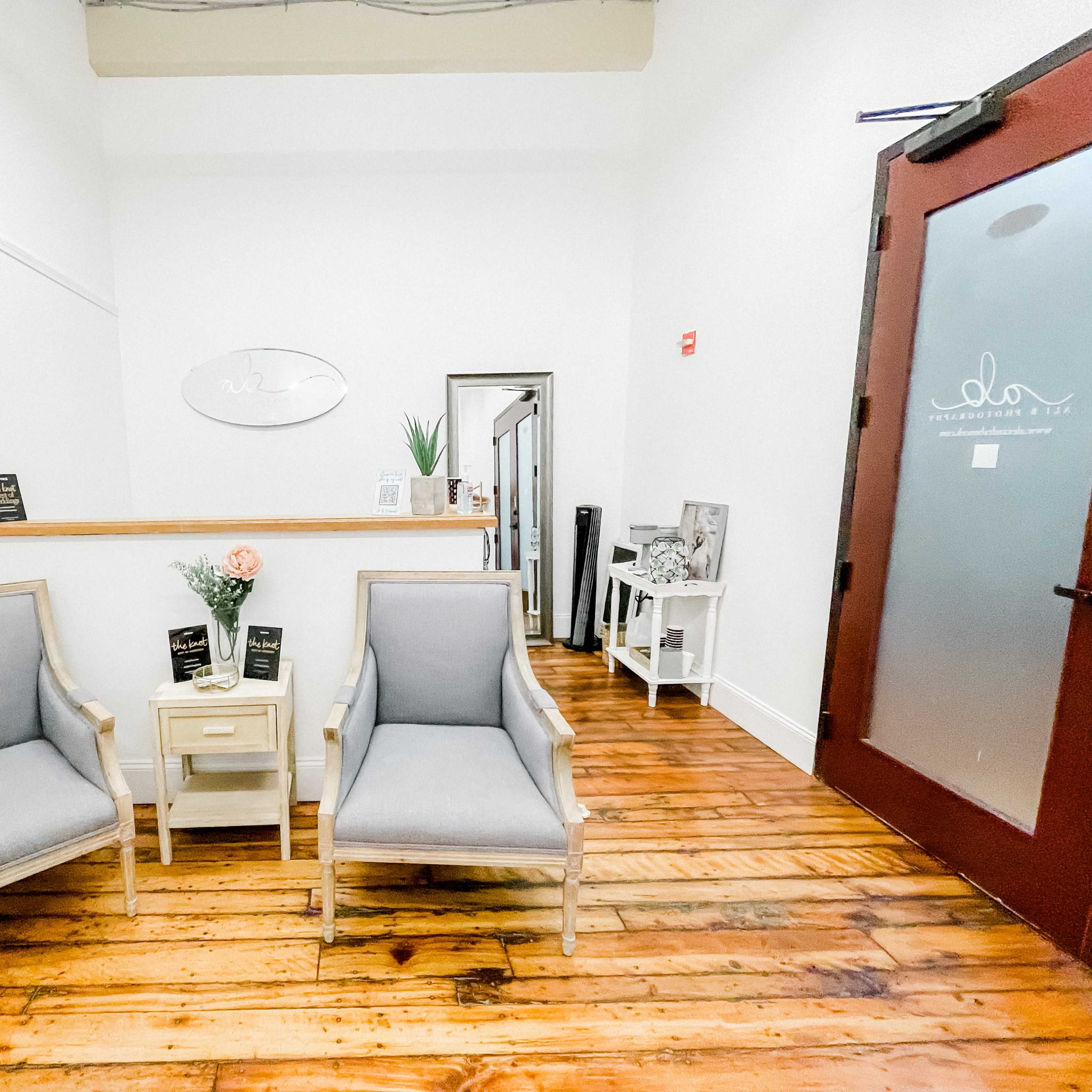 A waiting area with two gray chairs, a small table, and a reception desk against a white wall with wooden flooring.