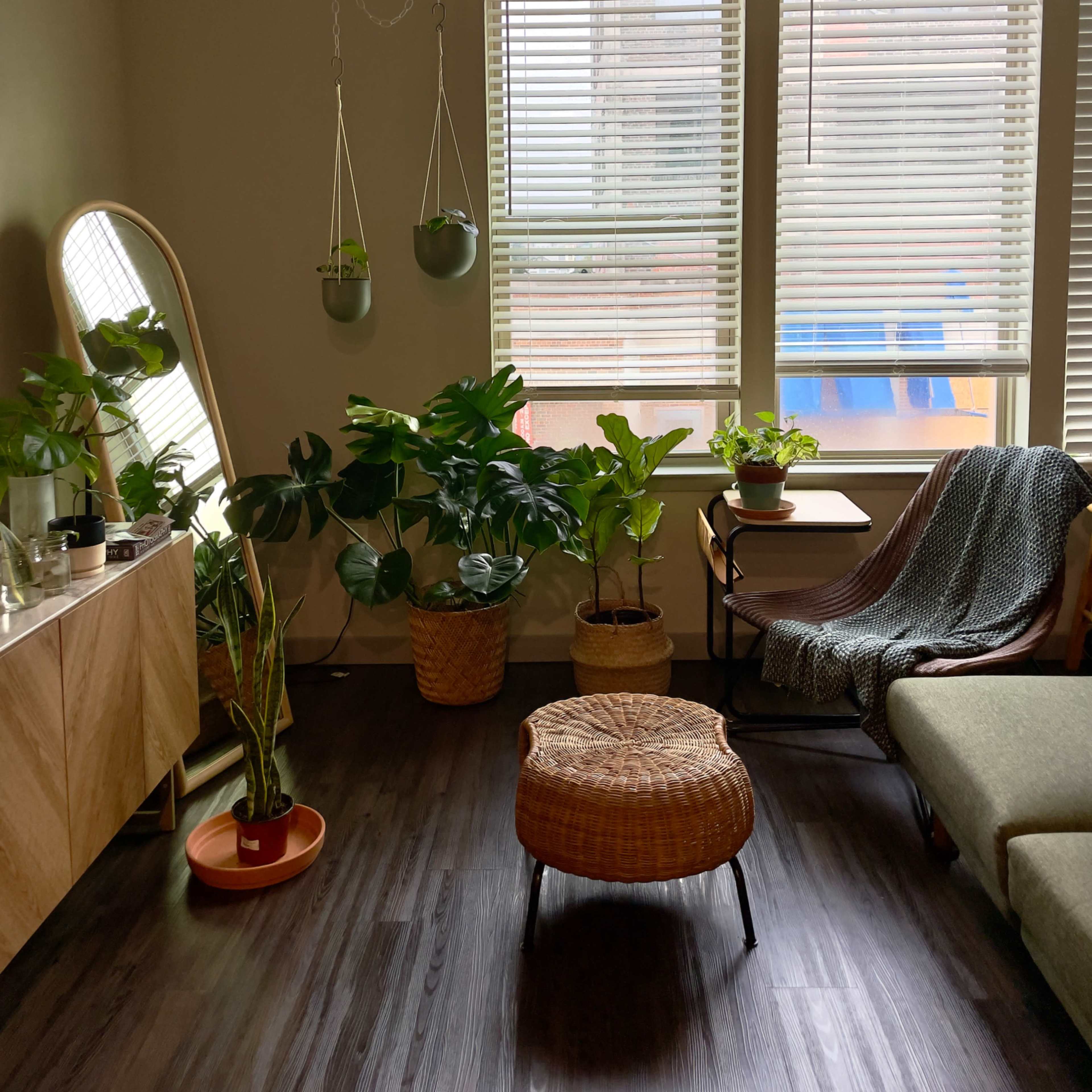 A cozy living room with large windows, several potted plants, a circular wicker ottoman, and a wooden sideboard.